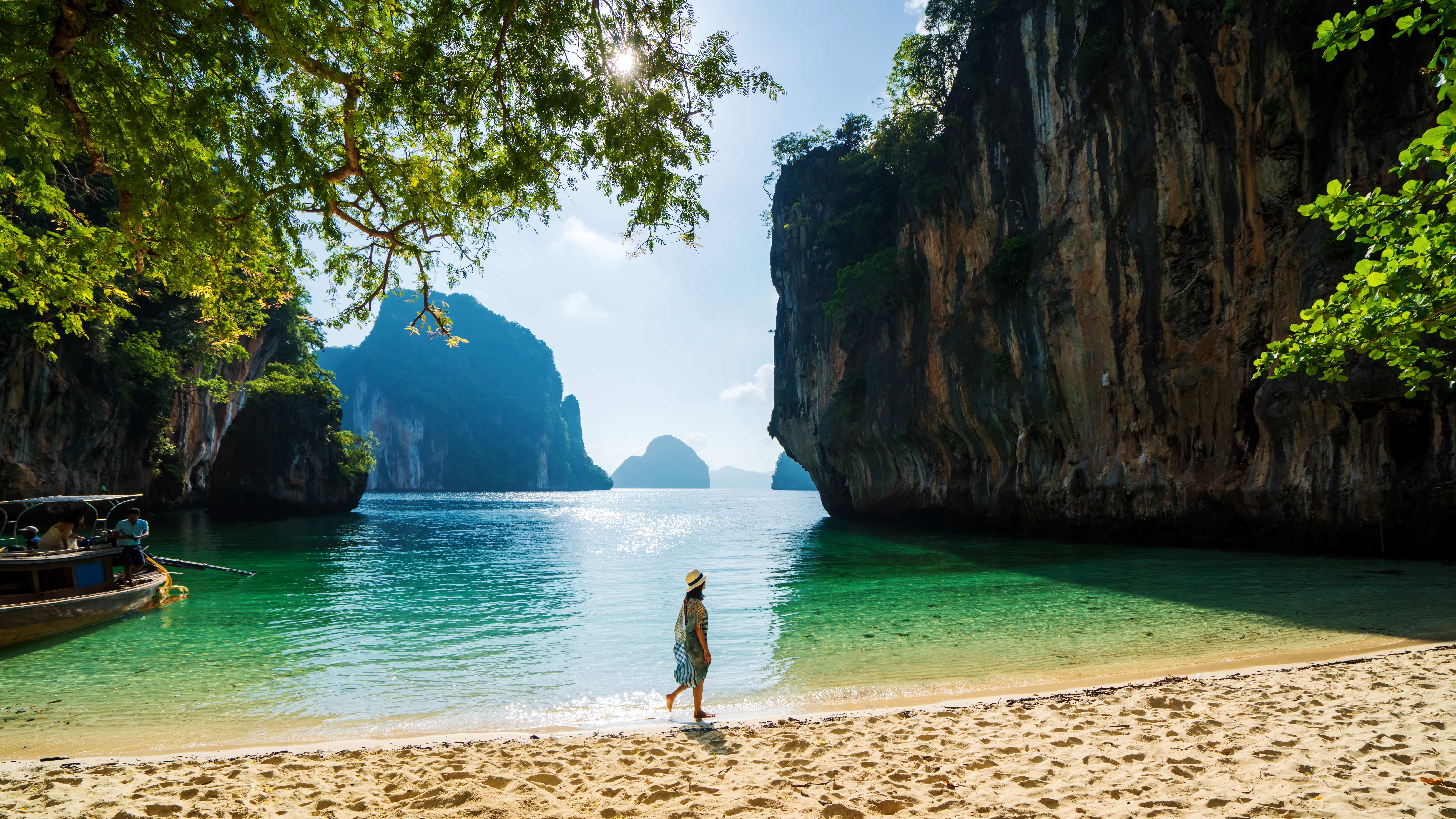 Person stands on a sandy beach facing turquoise water and towering cliffs.