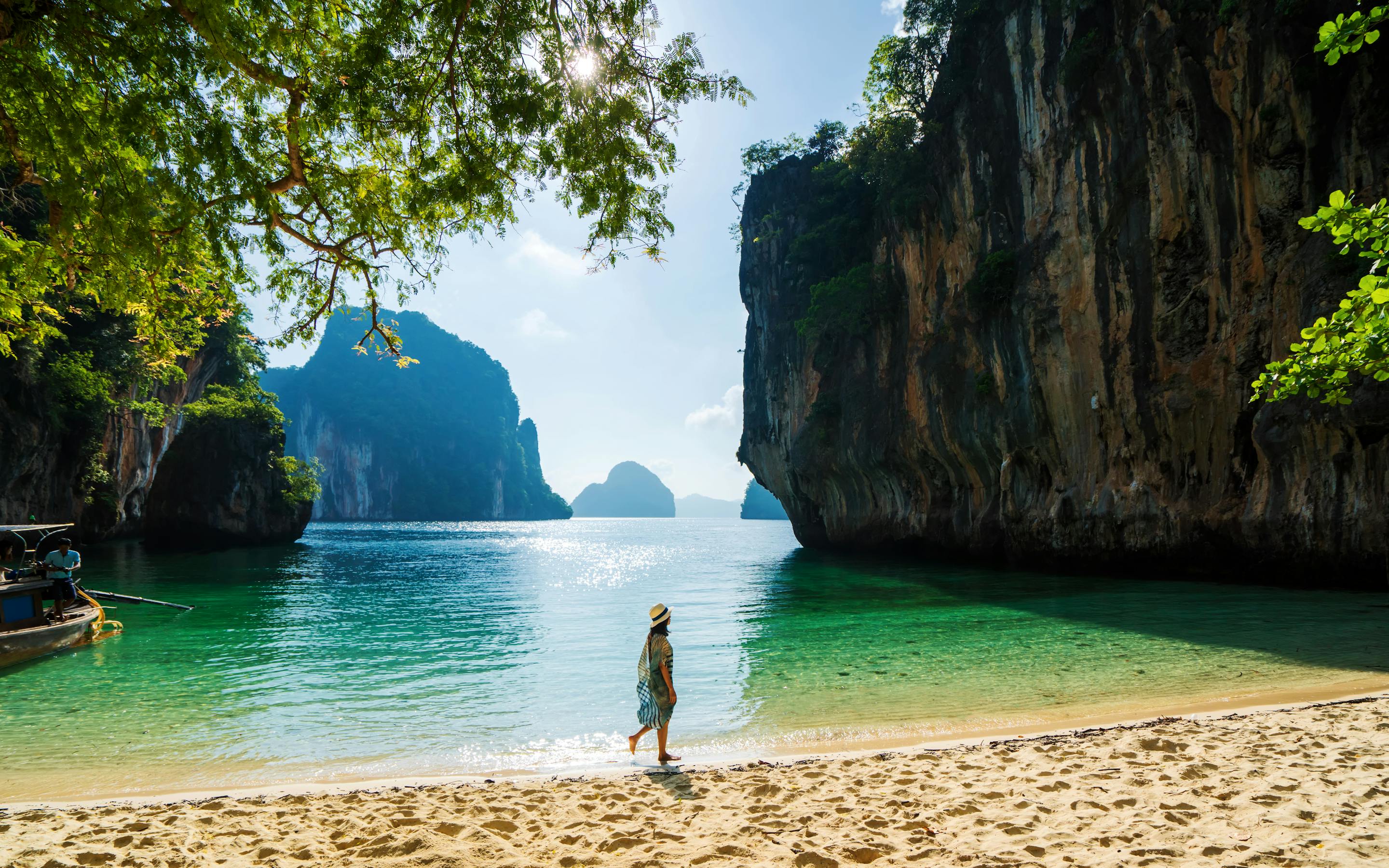 Person stands on a sandy beach facing turquoise water and towering cliffs.