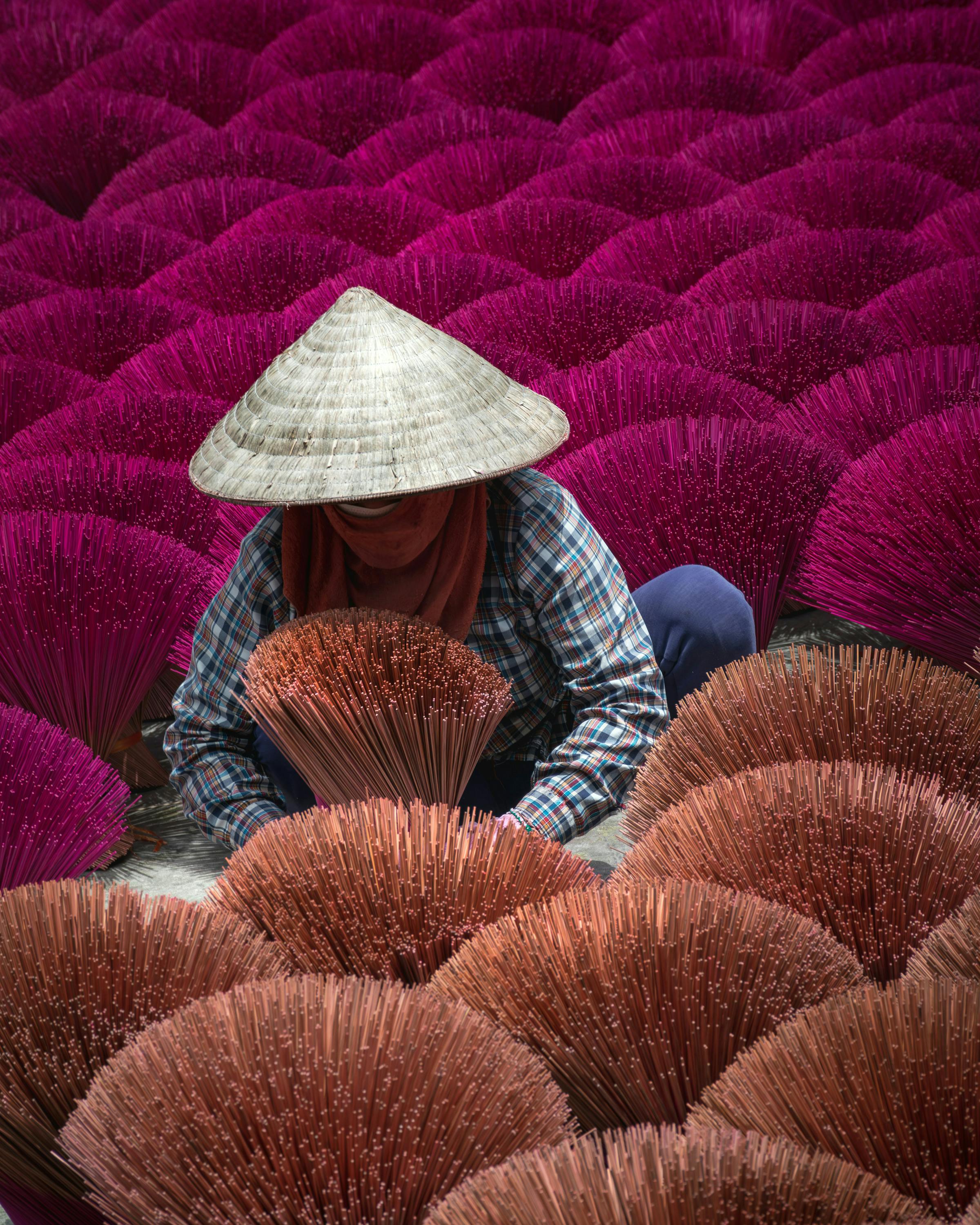 Person wearing a conical hat sits among rows of bright pink incense bundles.