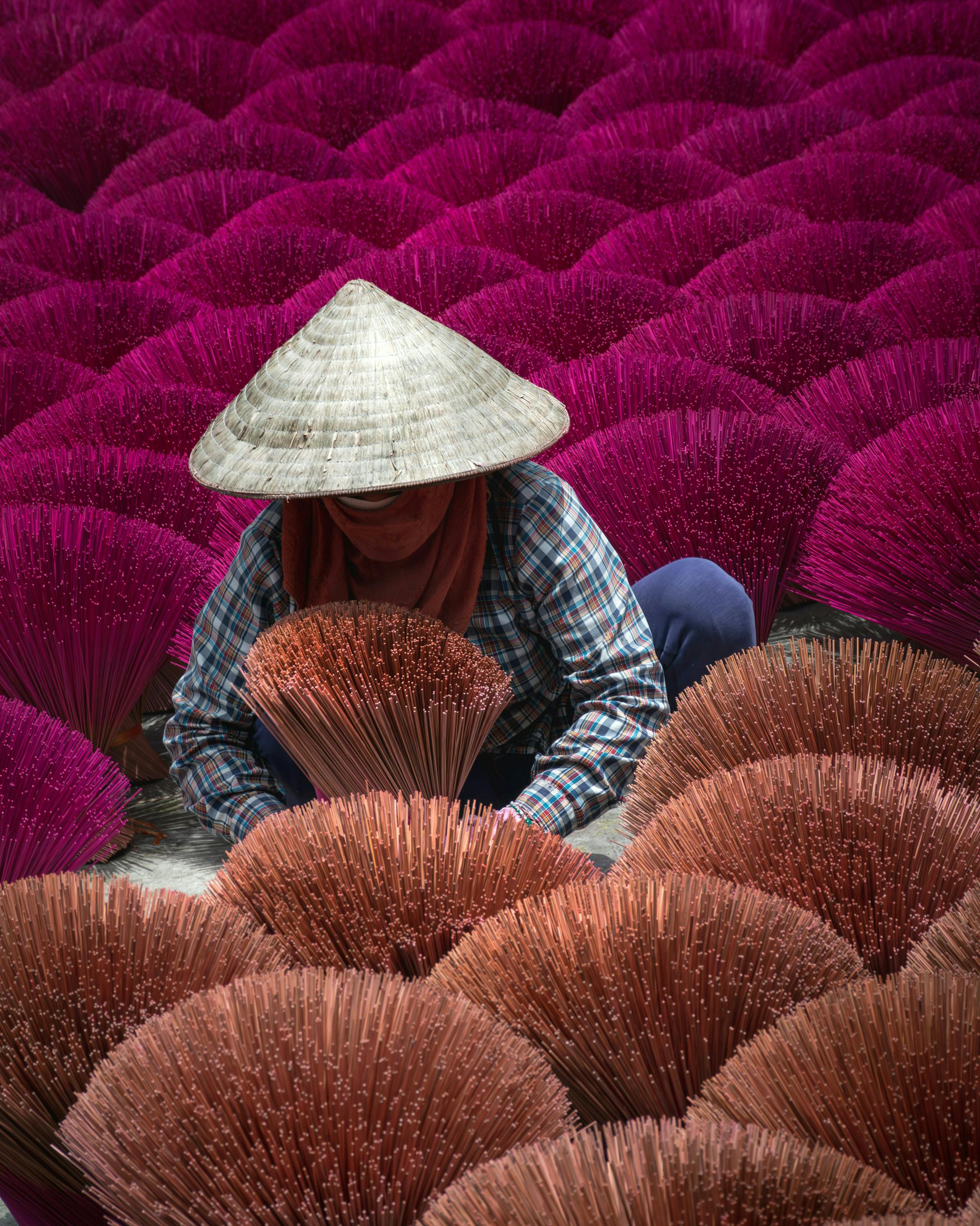 Person wearing a conical hat sits among rows of bright pink incense bundles.