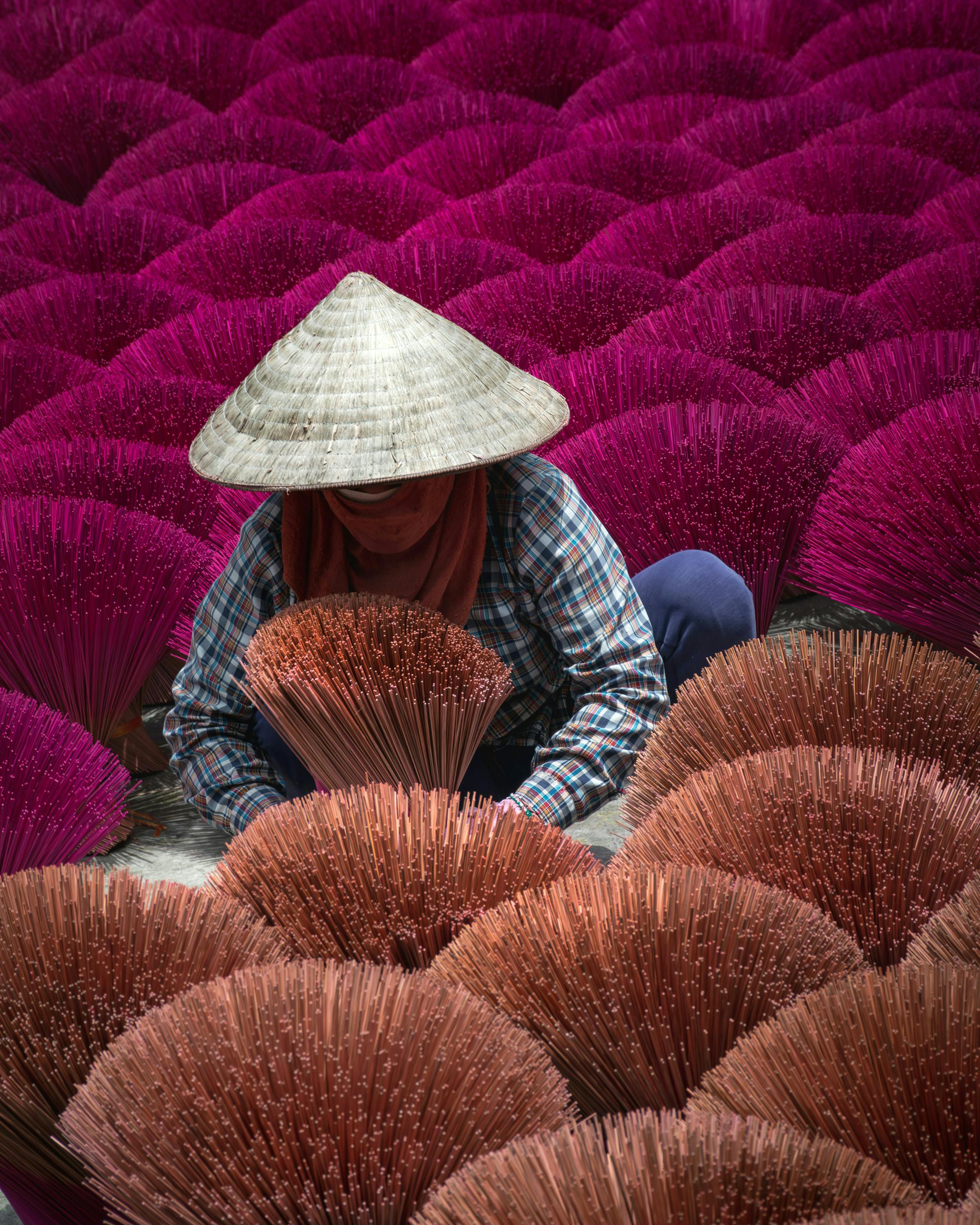 Person wearing a conical hat sits among rows of bright pink incense bundles.