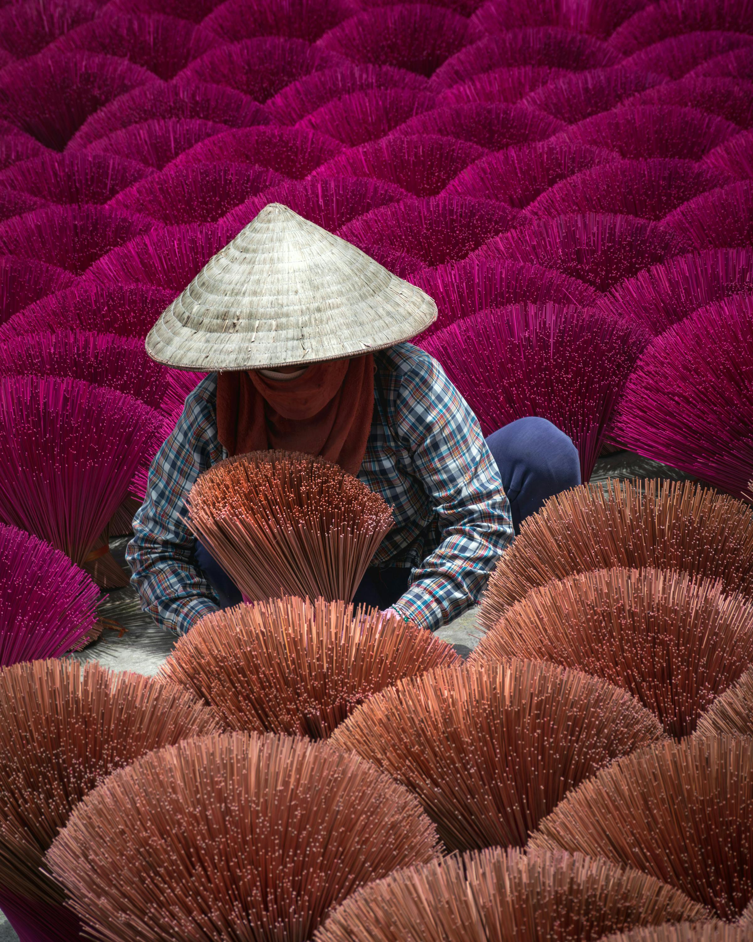 Person wearing a conical hat sits among rows of bright pink incense bundles.