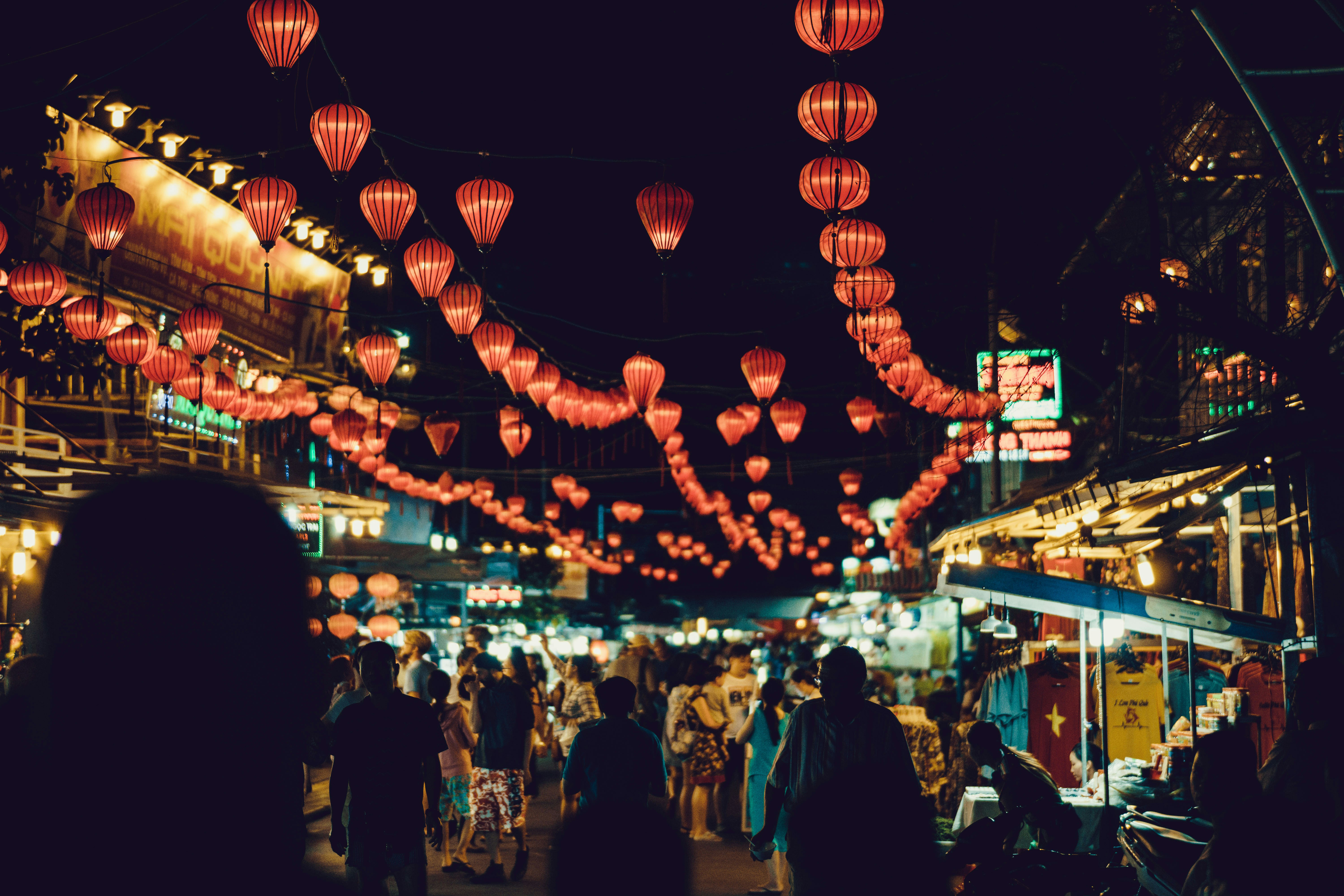 Crowded night street glows with hanging lanterns above market stalls.