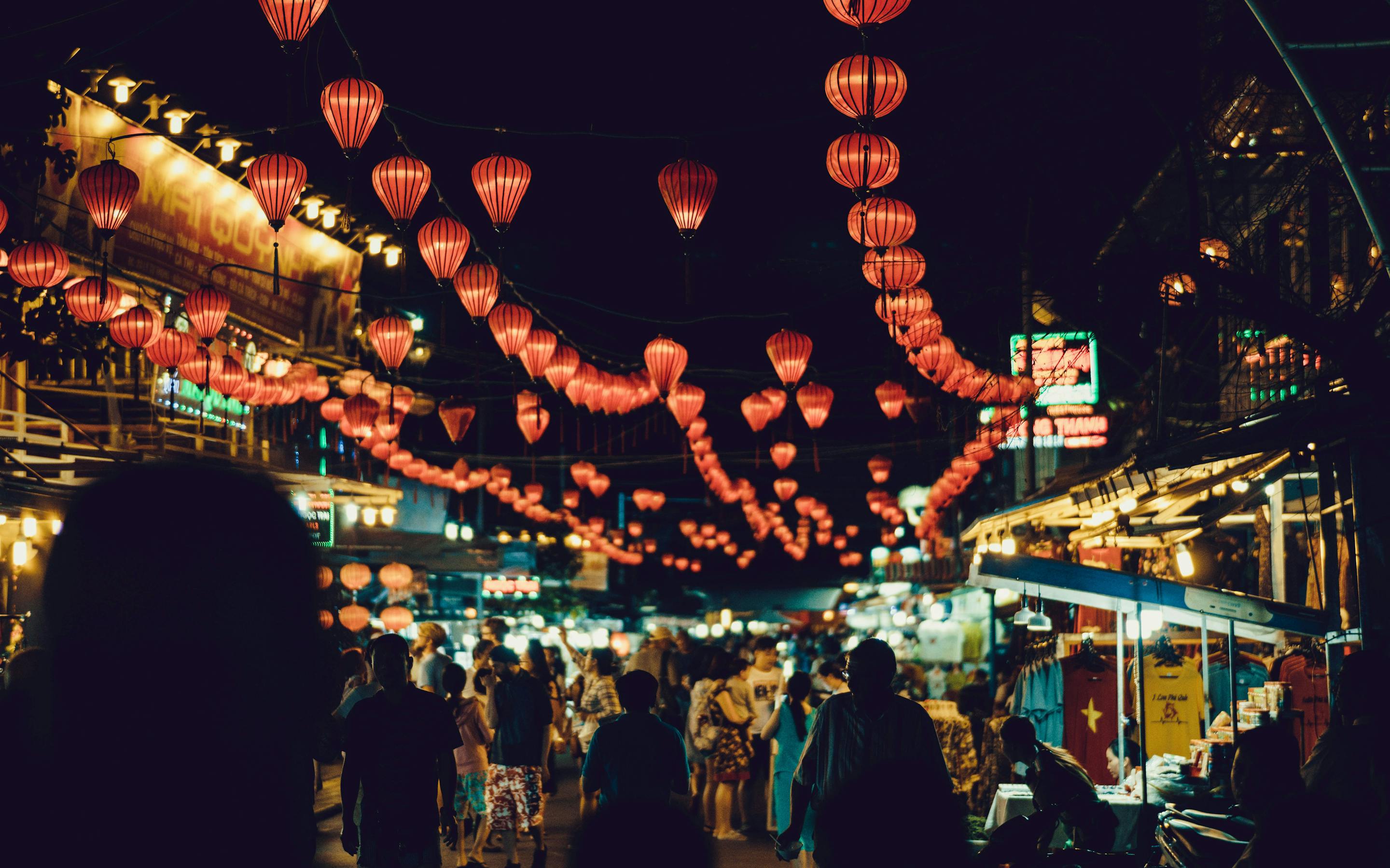Crowded night street glows with hanging lanterns above market stalls.