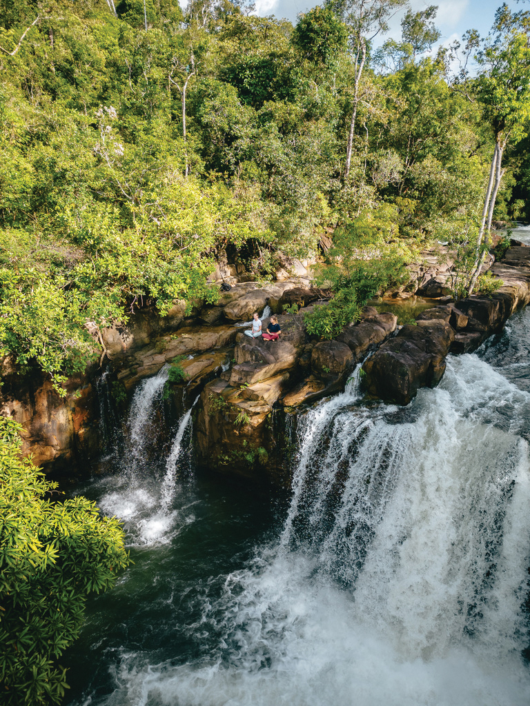 Person sits on rocks beside a waterfall cascading through lush forest.