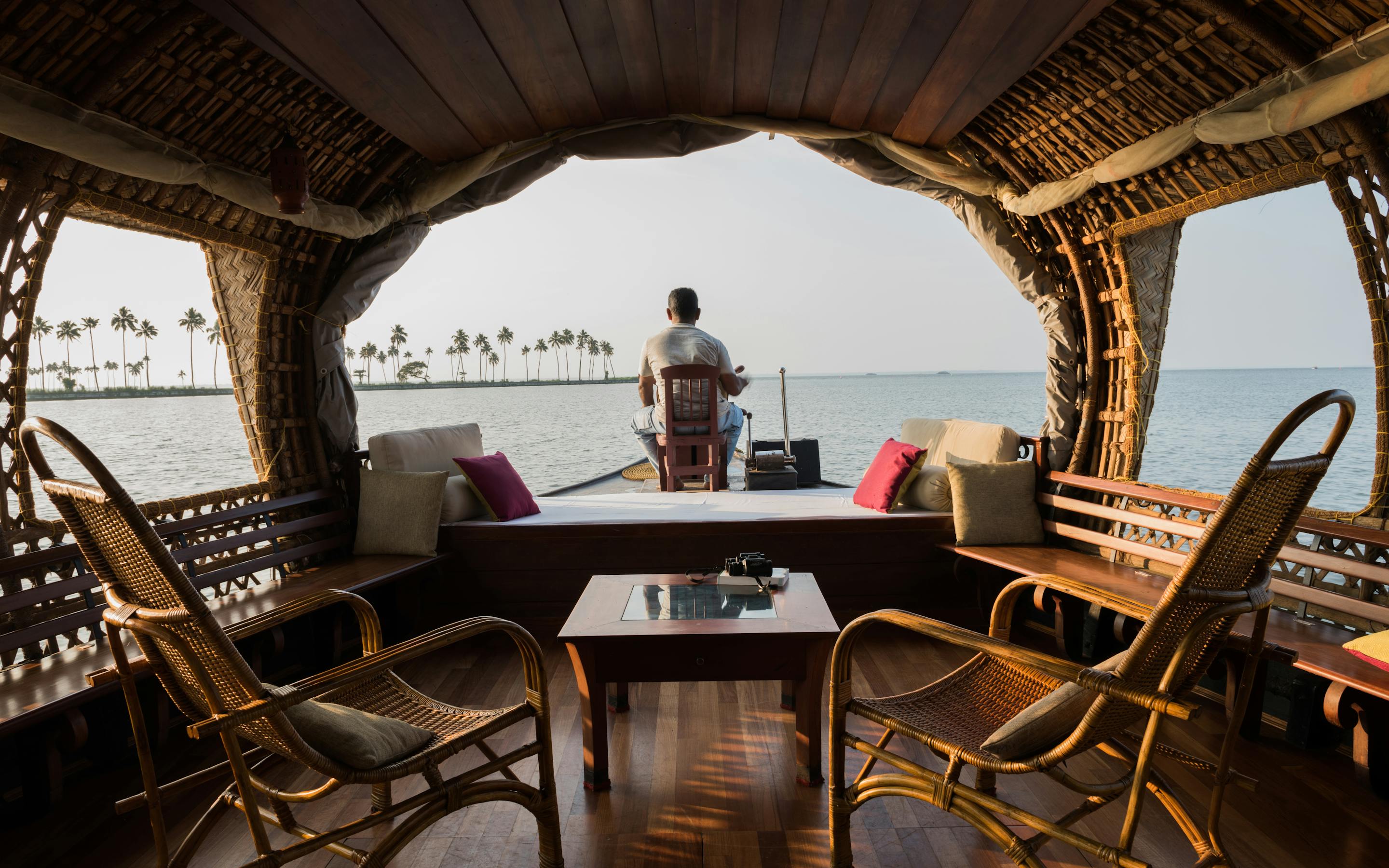 Person stands at the open front of a wooden houseboat with chairs and a table.