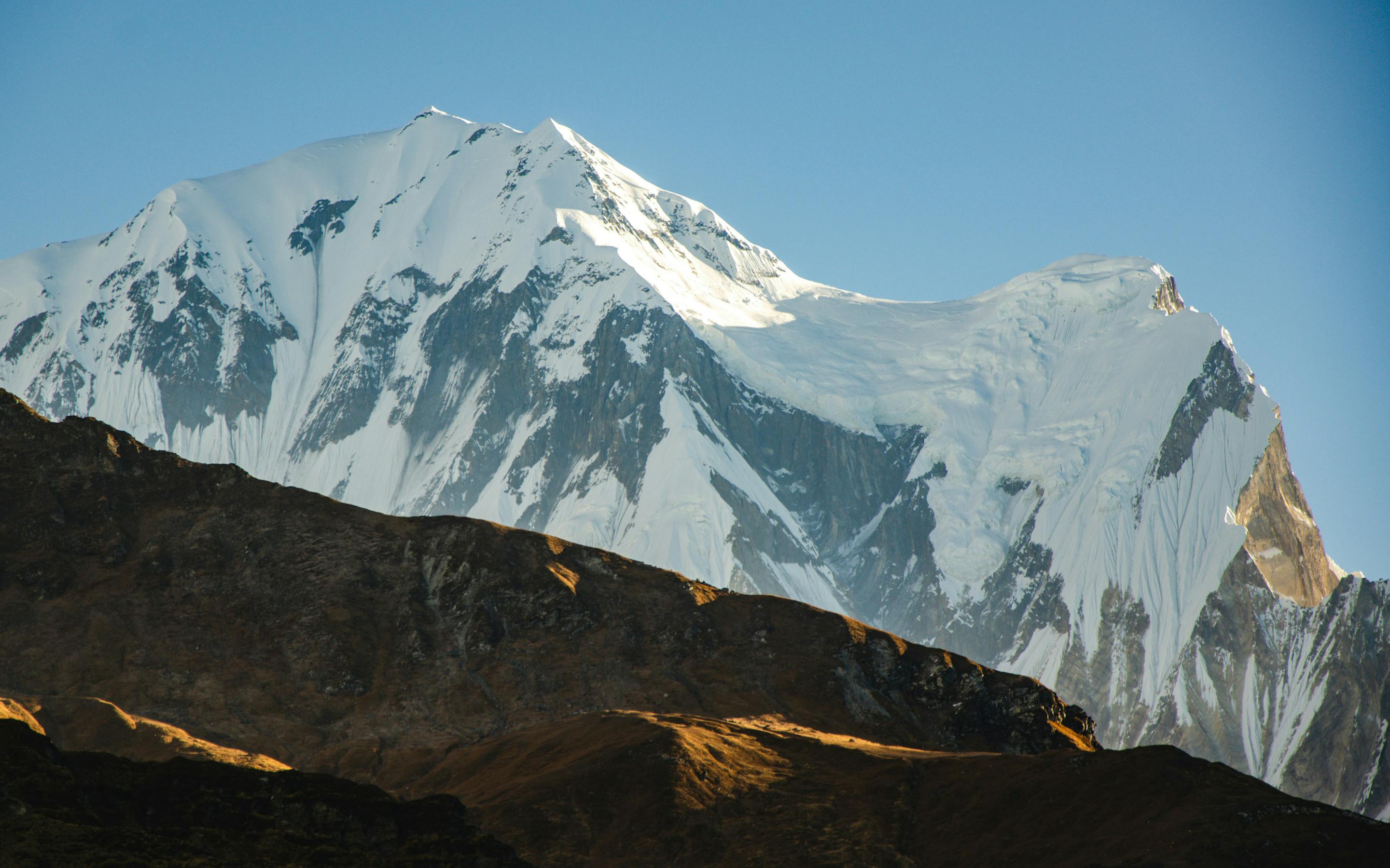 Snowy mountain peak rises above dark ridgelines under a clear blue sky.