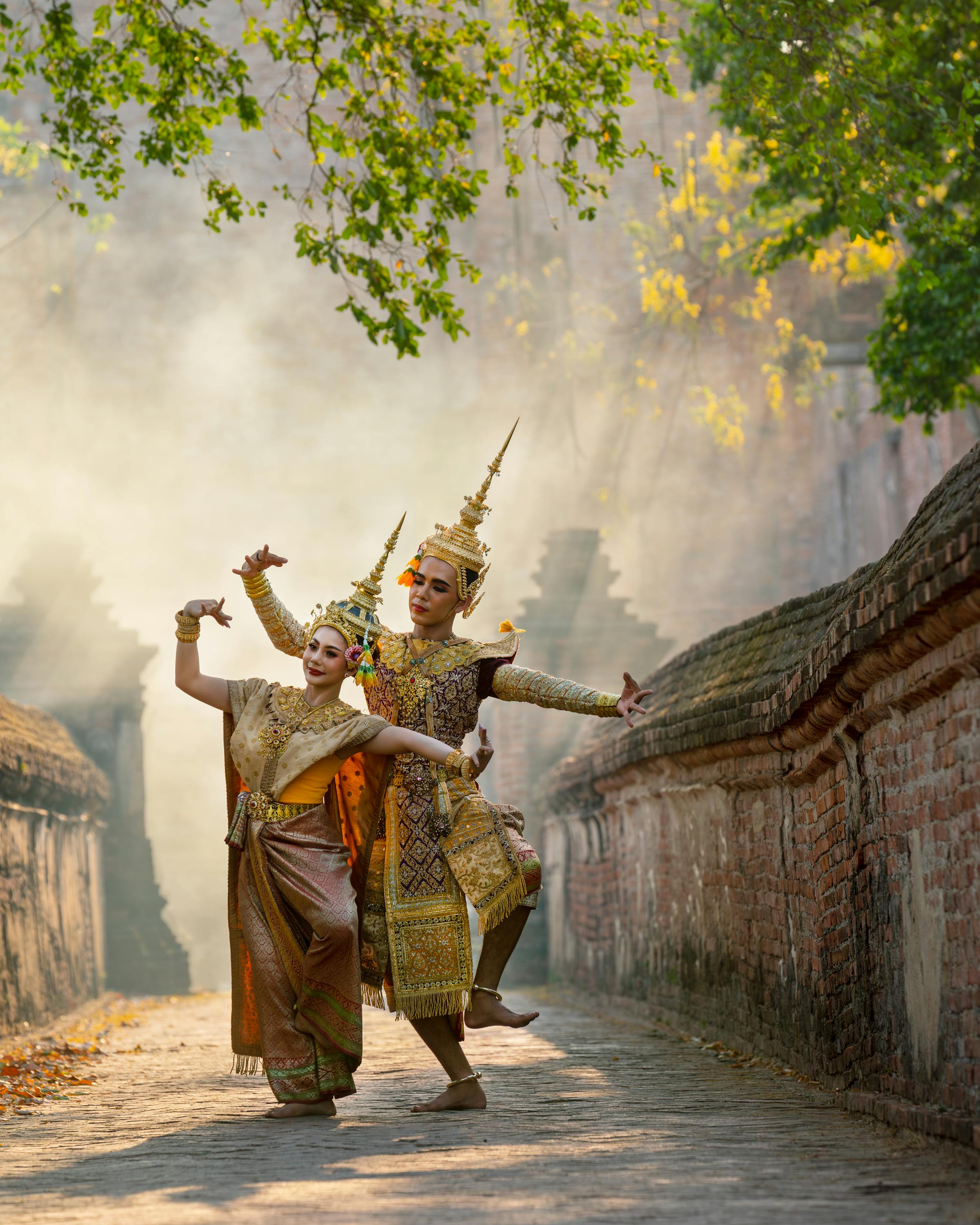 Two performers in ornate costumes dance in a sunlit courtyard with smoke in the background.
