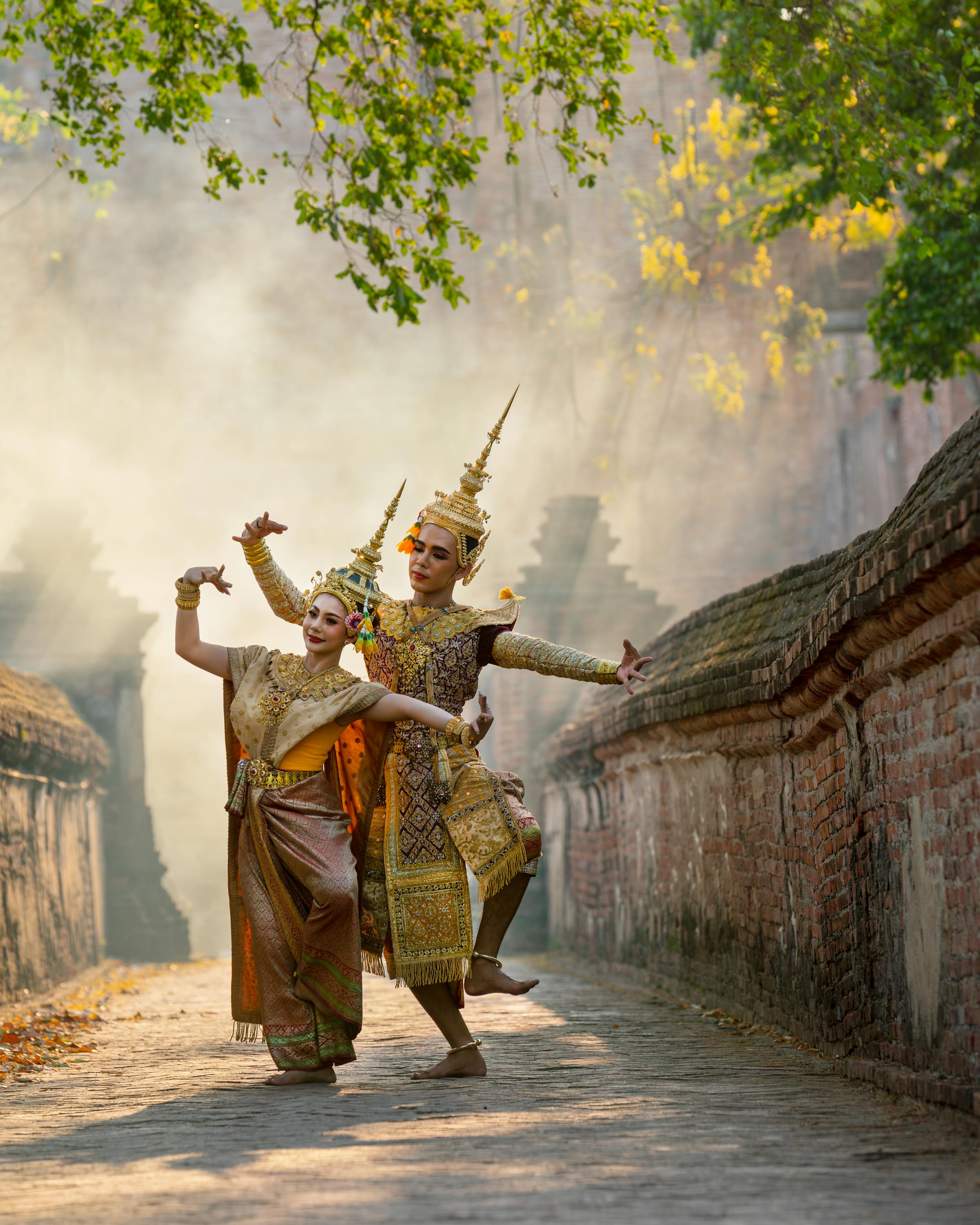 Two performers in ornate costumes dance in a sunlit courtyard with smoke in the background.
