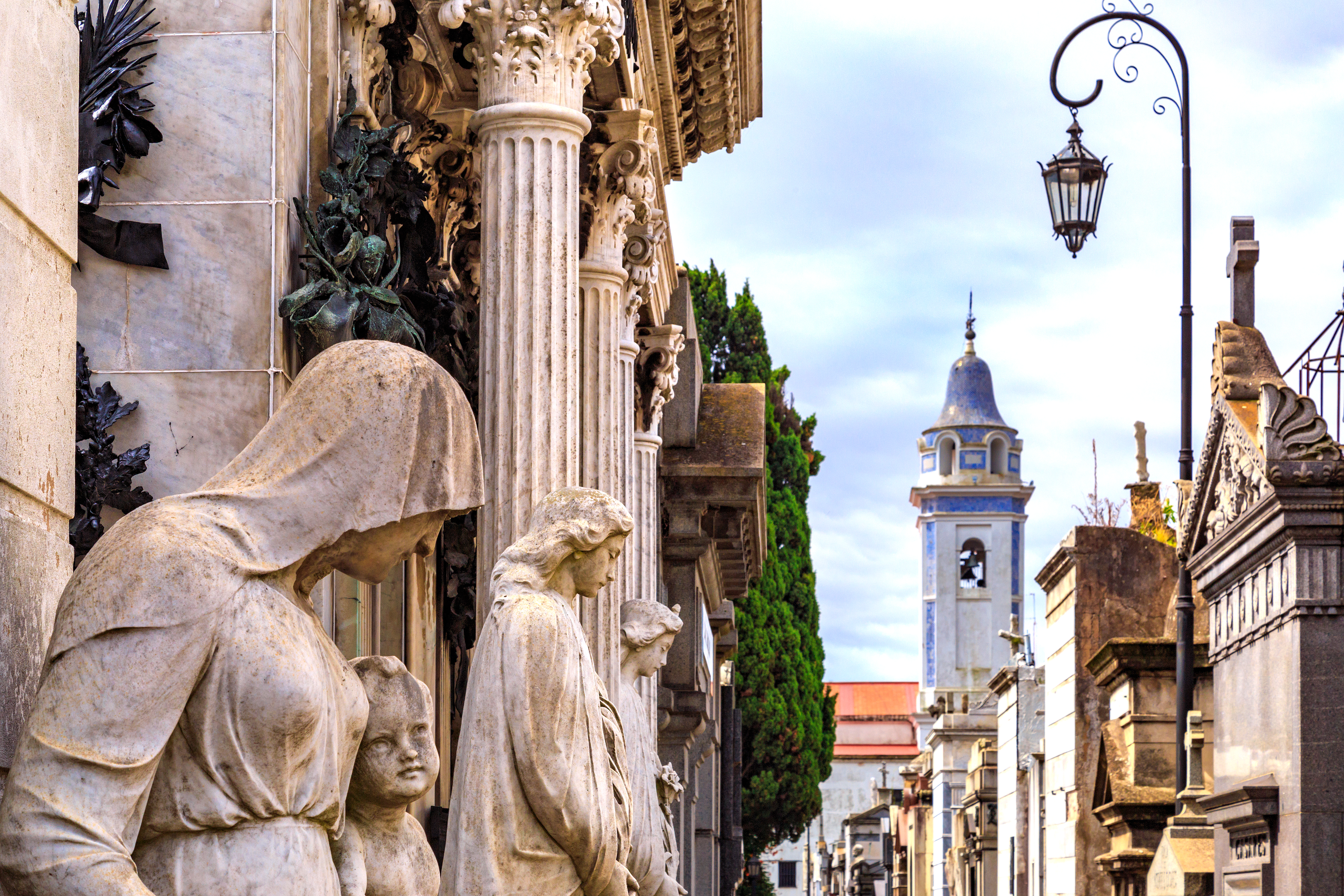 Stone statues line a narrow street leading toward a church tower under a clear sky.