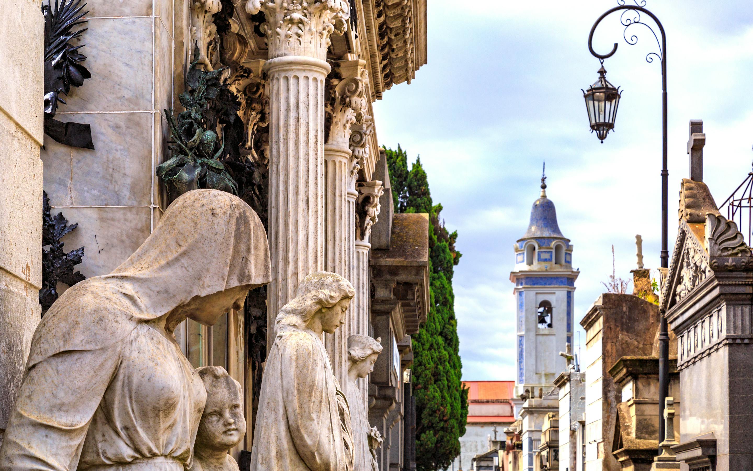 Stone statues line a narrow street leading toward a church tower under a clear sky.