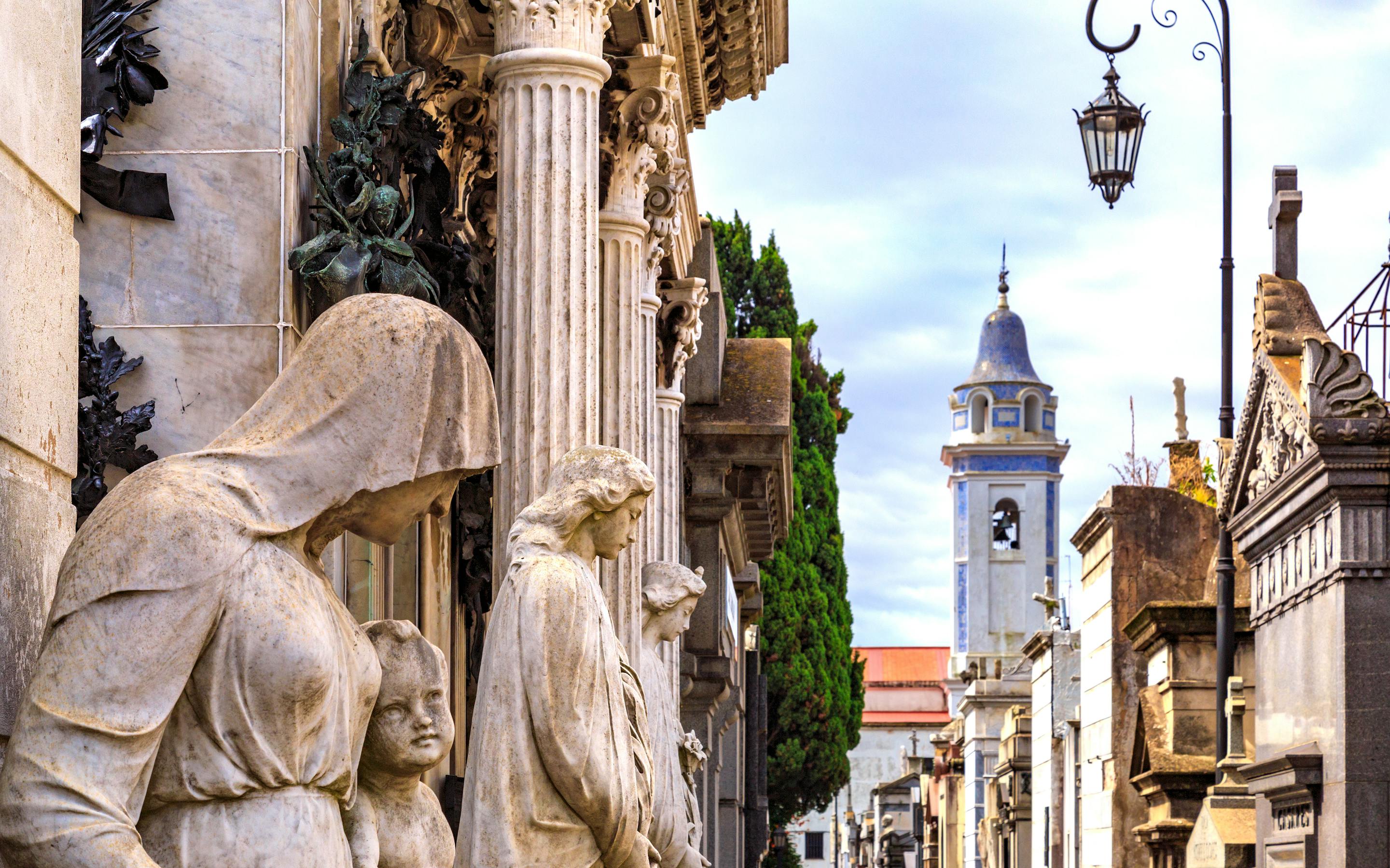 Stone statues line a narrow street leading toward a church tower under a clear sky.
