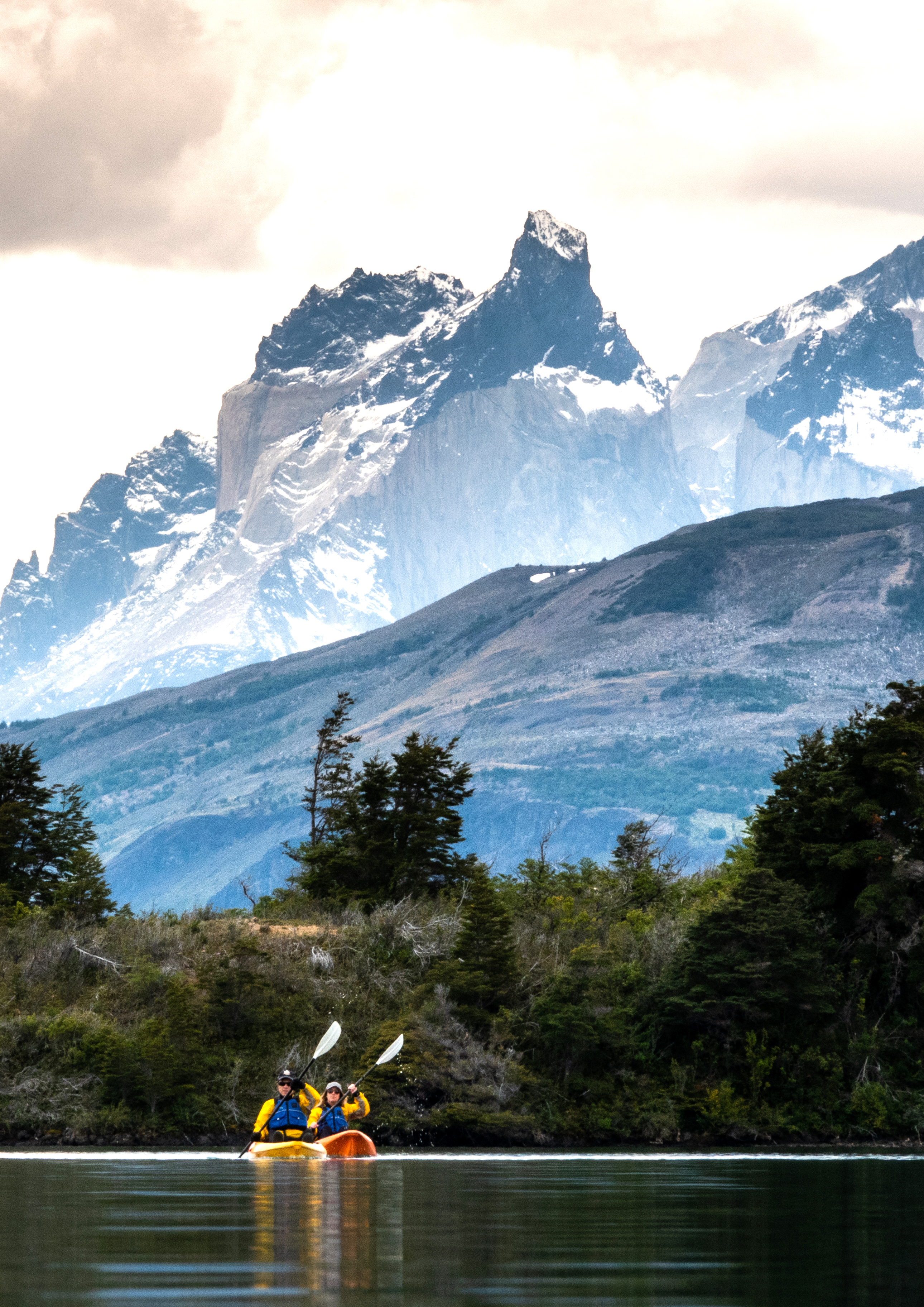 Two people kayak on a calm lake with dramatic mountain peaks and cloudy skies beyond.