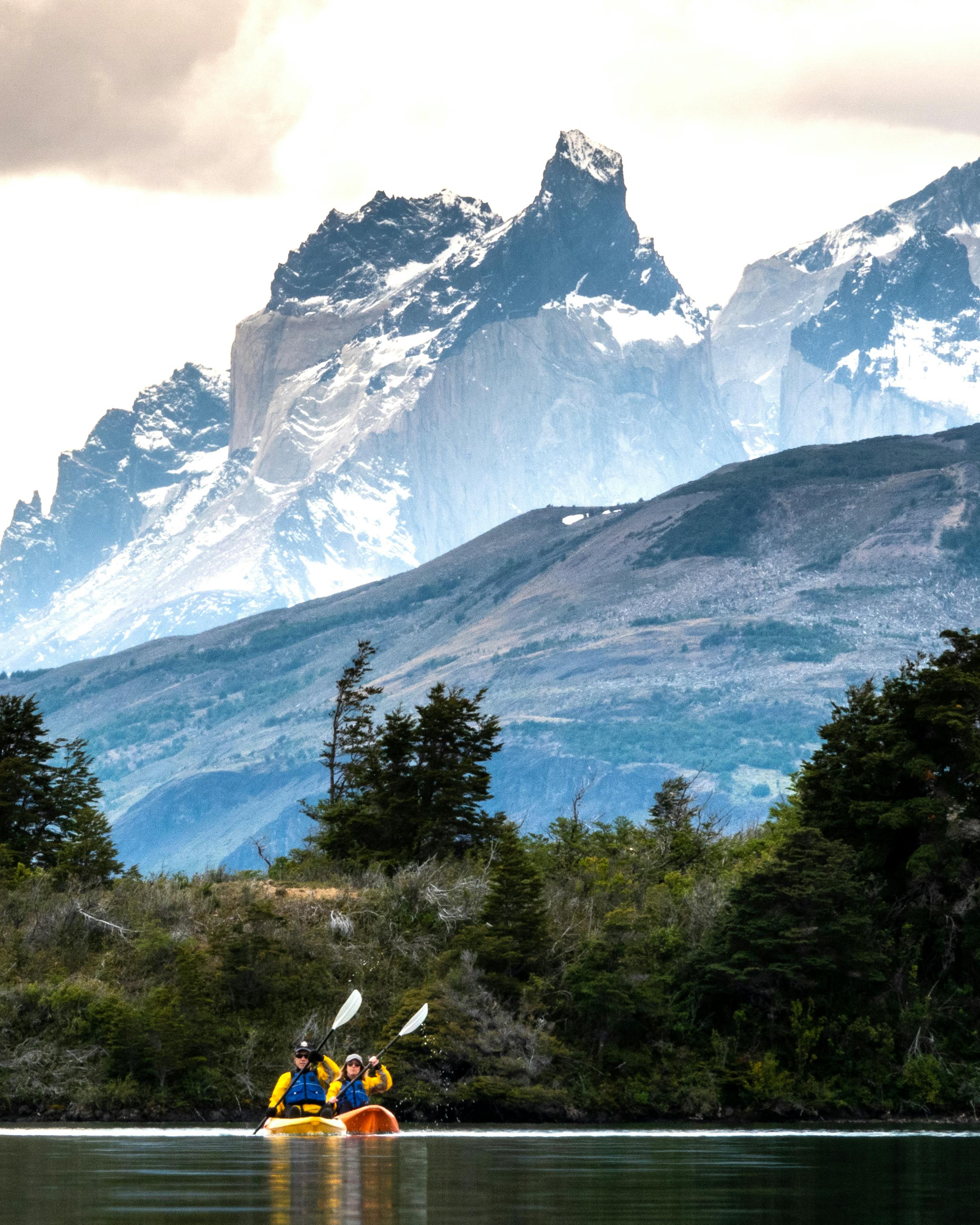 Two people kayak on a calm lake with dramatic mountain peaks and cloudy skies beyond.