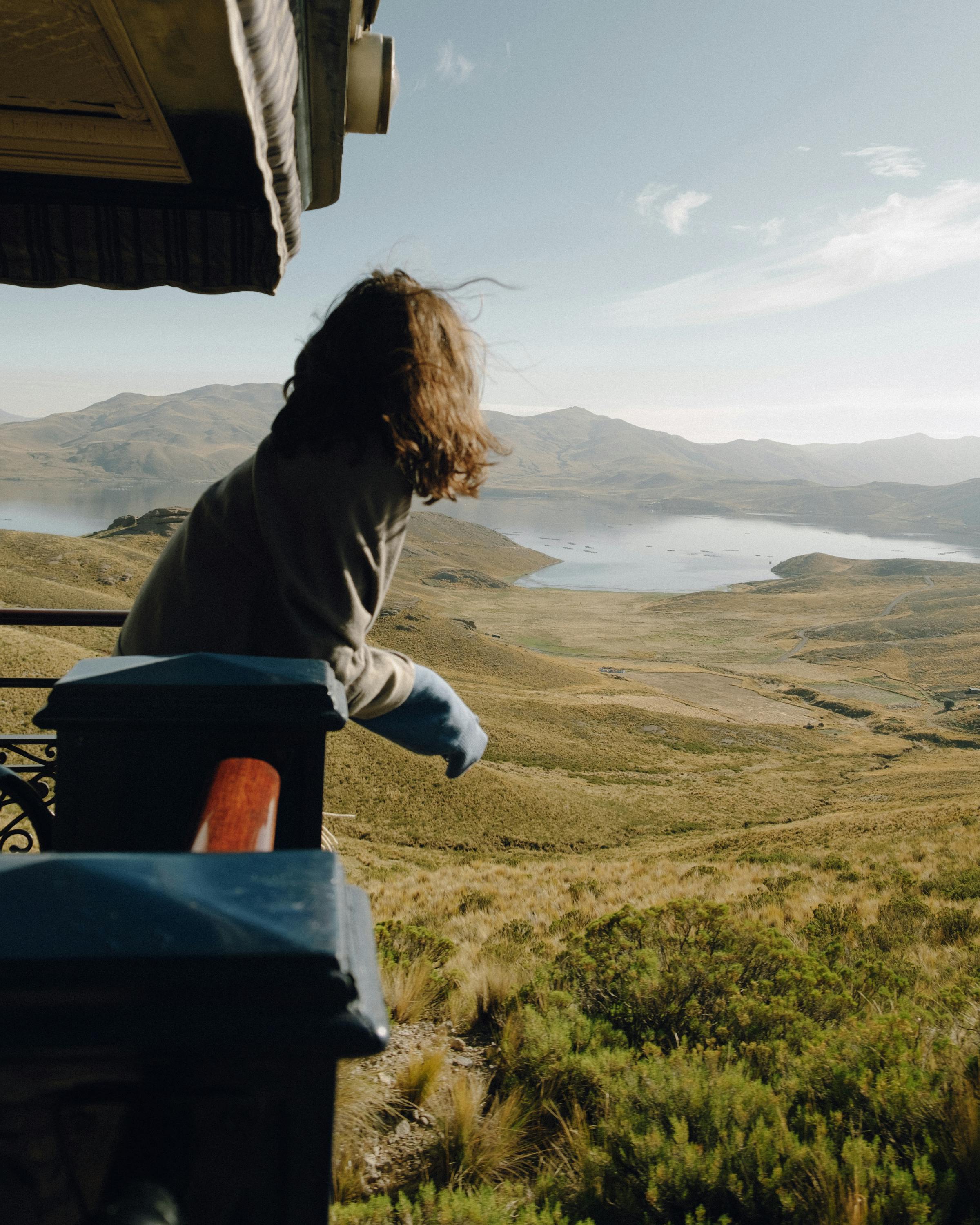 Person leans from a train window looking out over rolling hills and distant water.