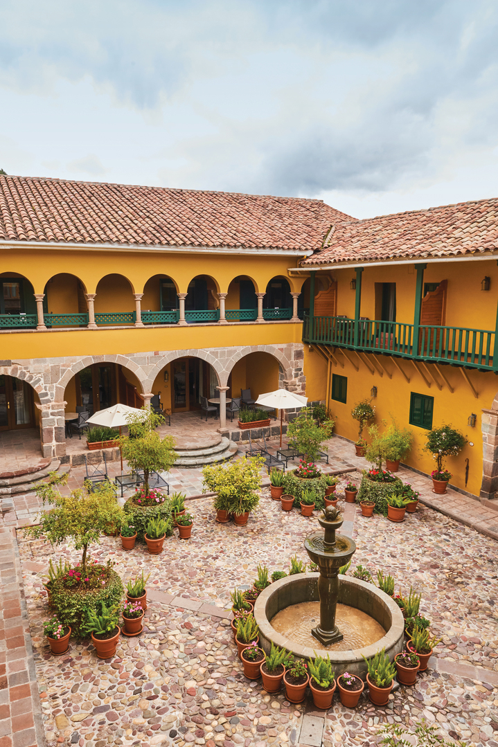 Sunlit courtyard with a central fountain and potted plants surrounded by yellow arches.