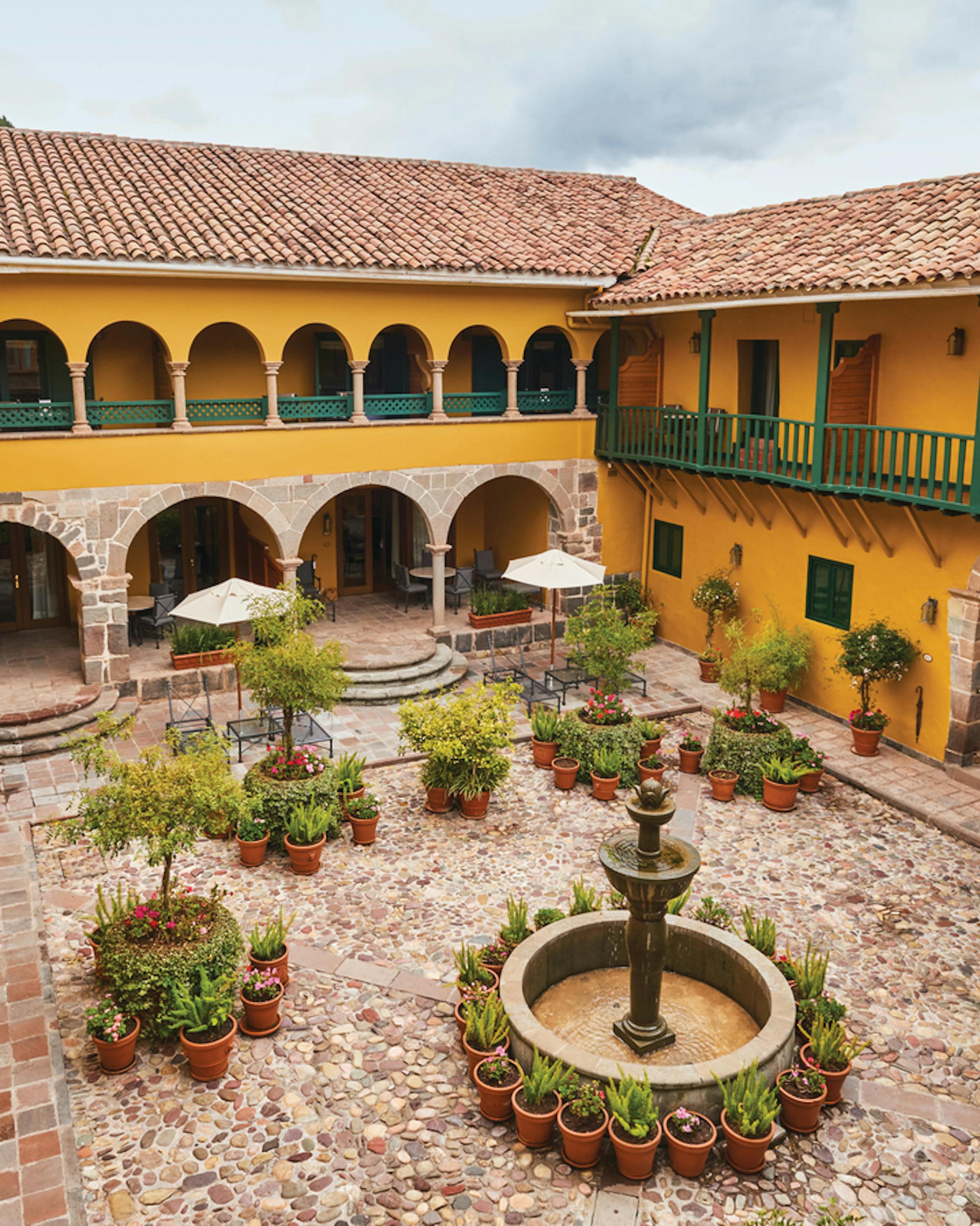 Sunlit courtyard with a central fountain and potted plants surrounded by yellow arches.