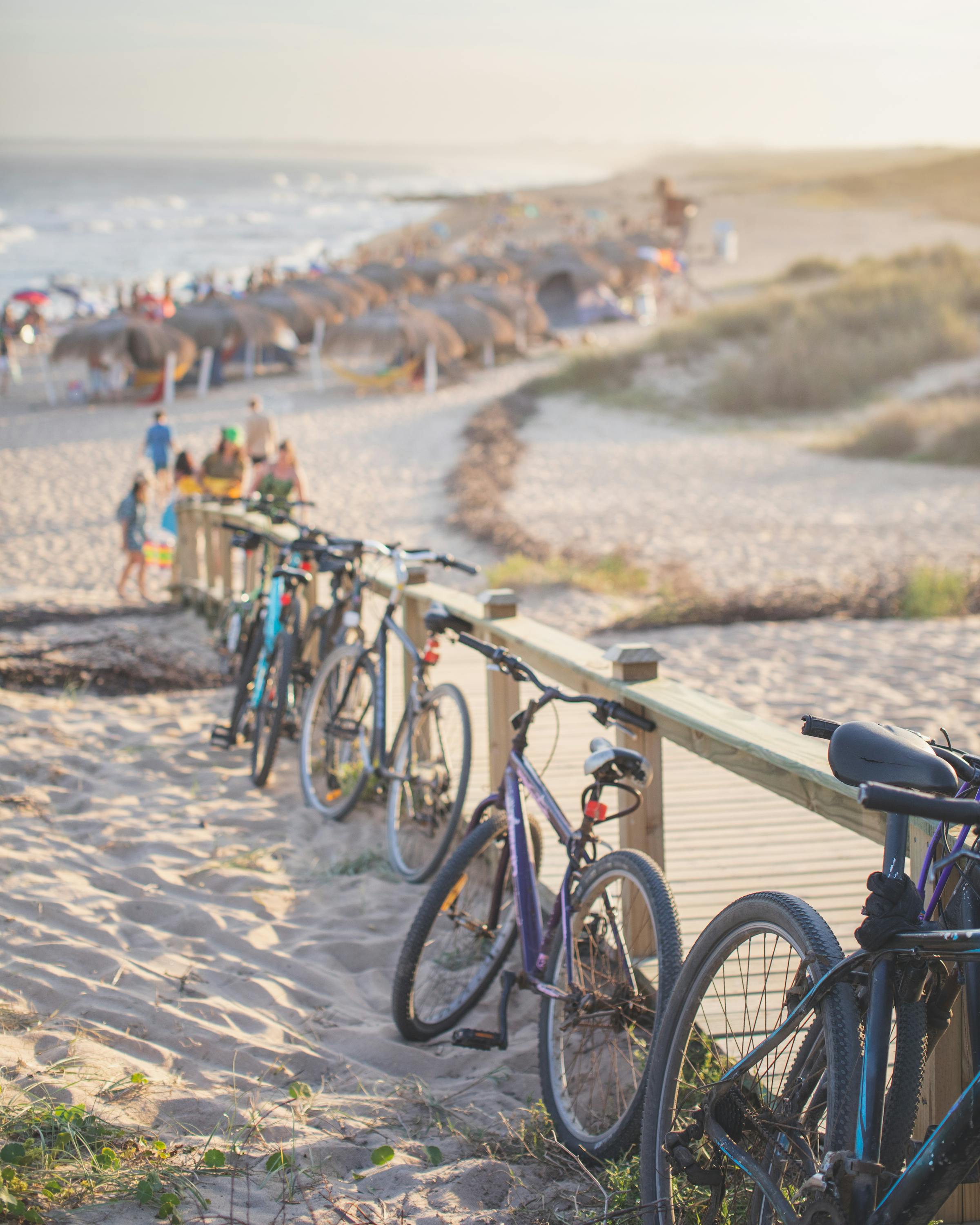 Bicycles line a sandy path overlooking a busy beach and ocean under hazy sunlight.