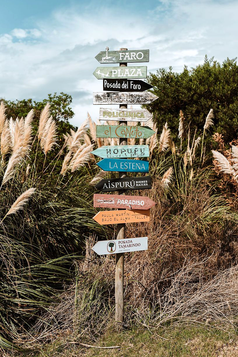 Weathered signpost with many directional signs stands in sandy dunes with grasses.