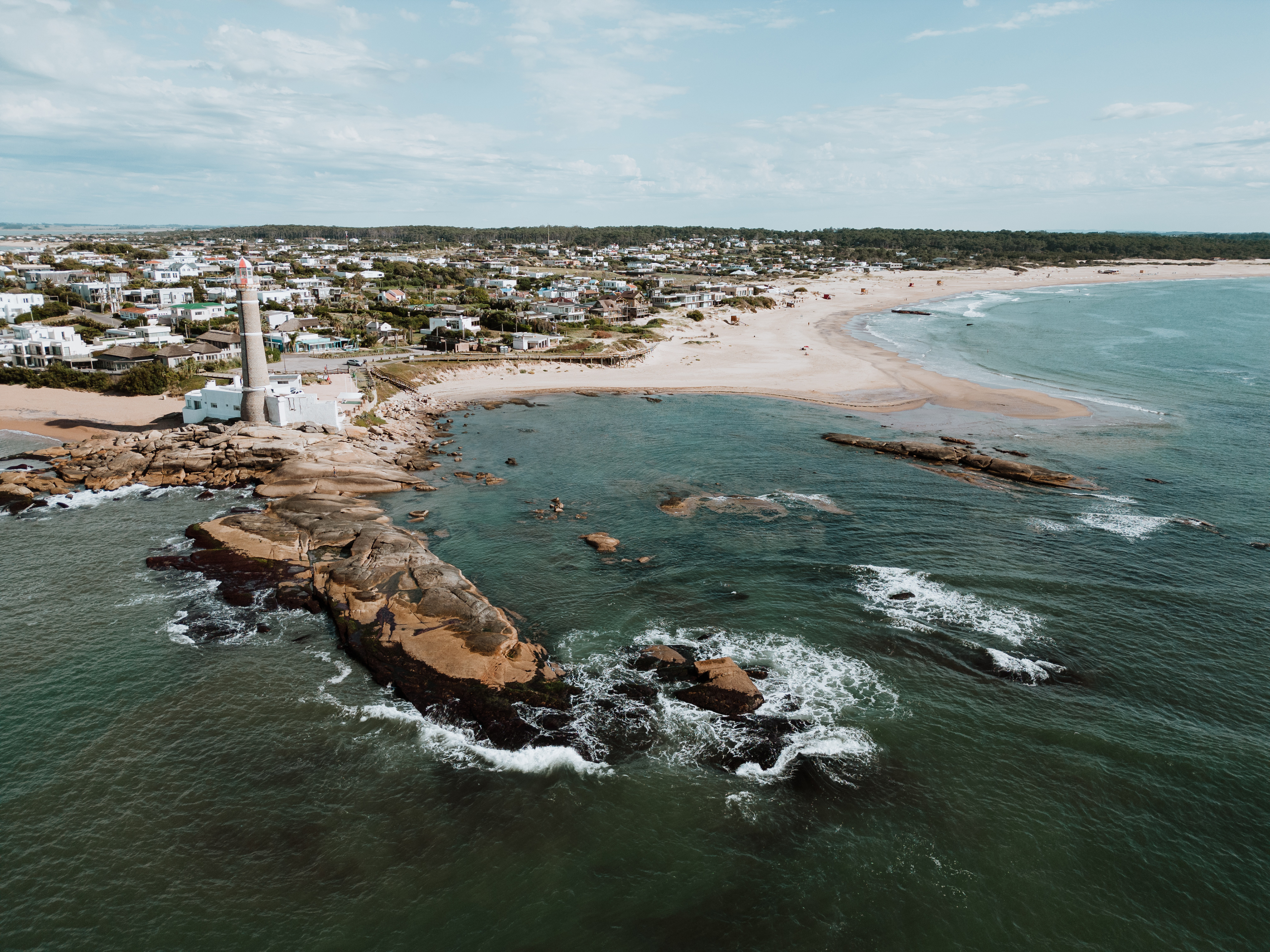 Aerial view of a rocky point and lighthouse beside a sandy beach and coastal town.