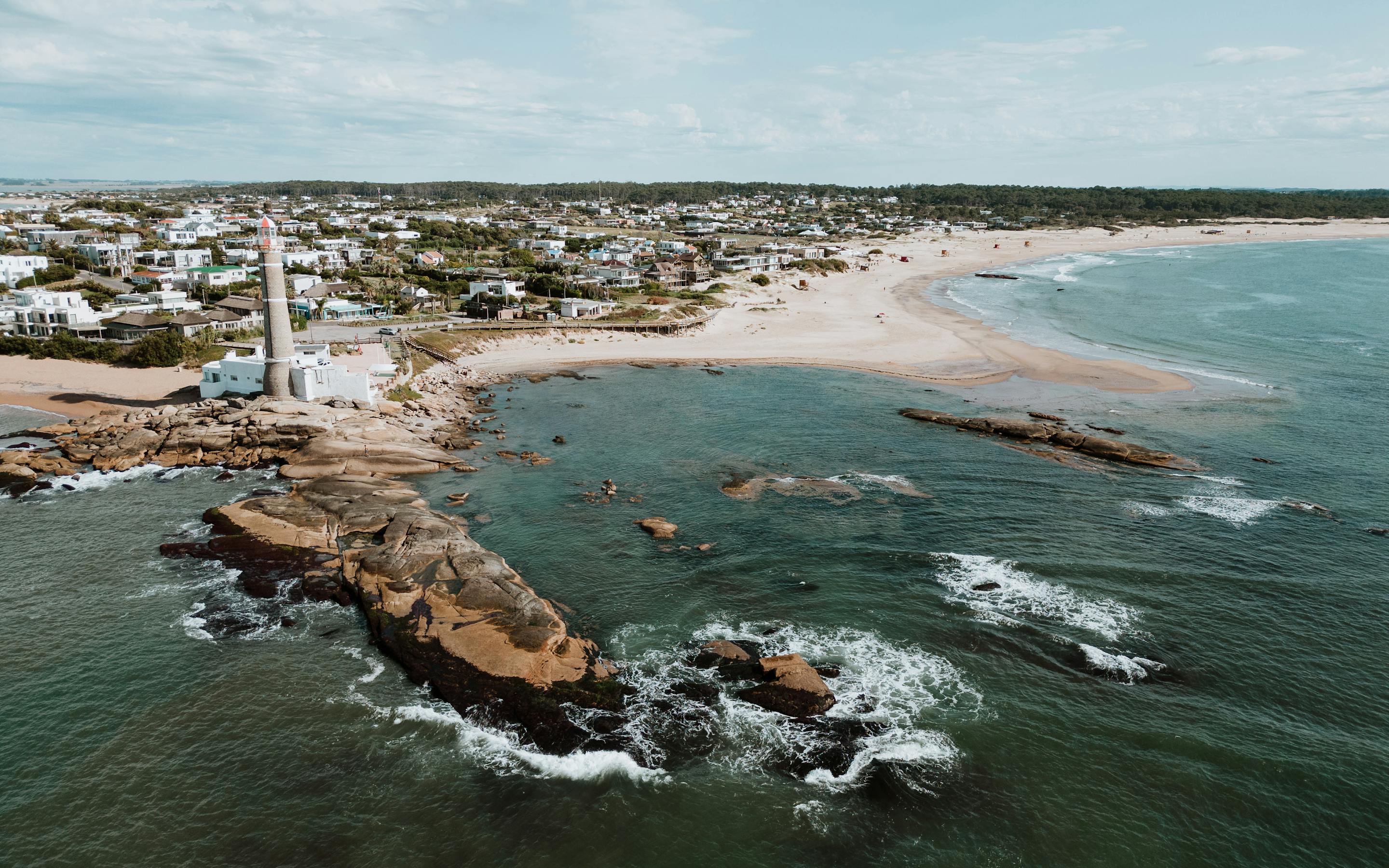 Aerial view of a rocky point and lighthouse beside a sandy beach and coastal town.