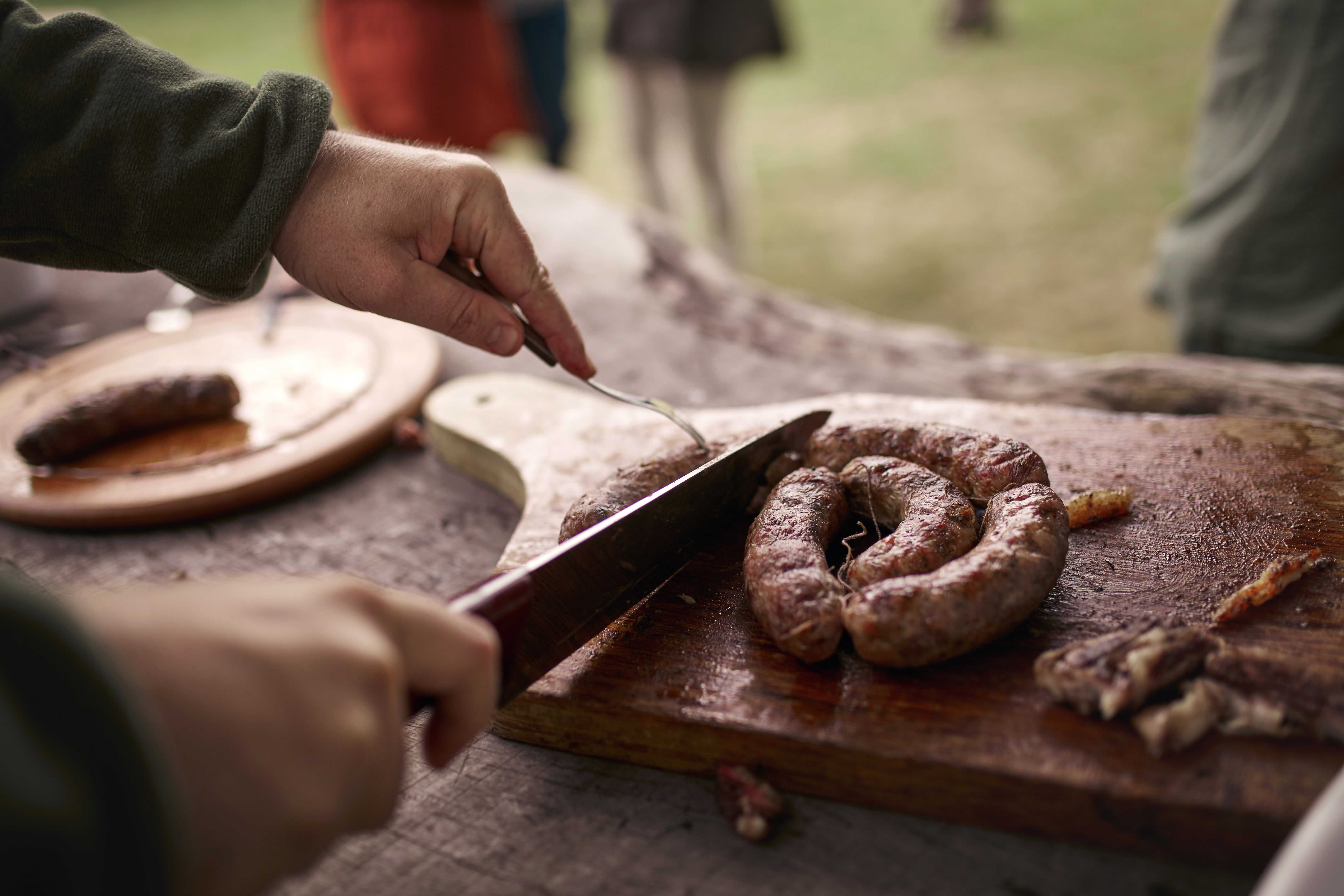 Hands slice sausage on a wooden board beside plates at an outdoor table.