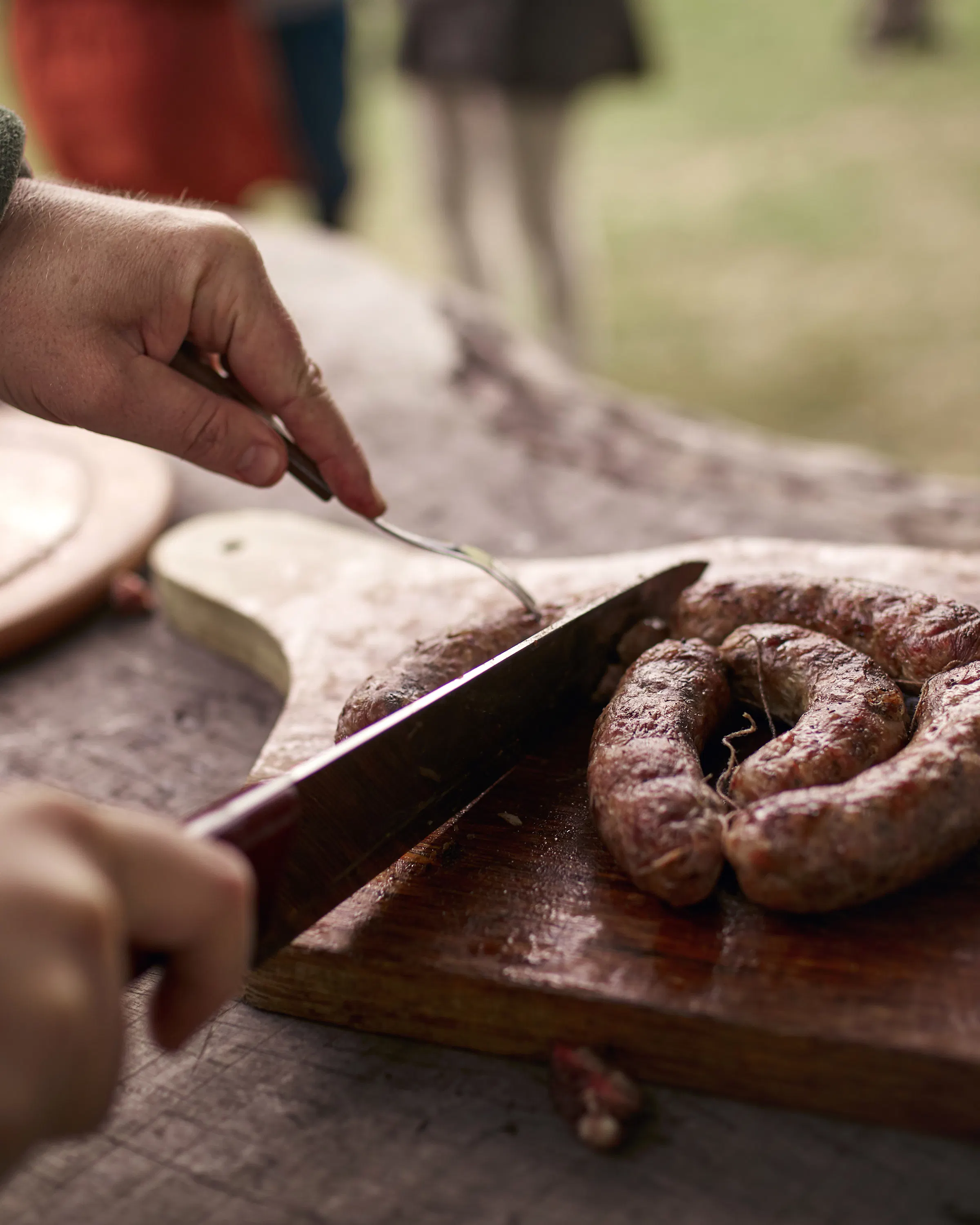 Hands slice sausage on a wooden board beside plates at an outdoor table.