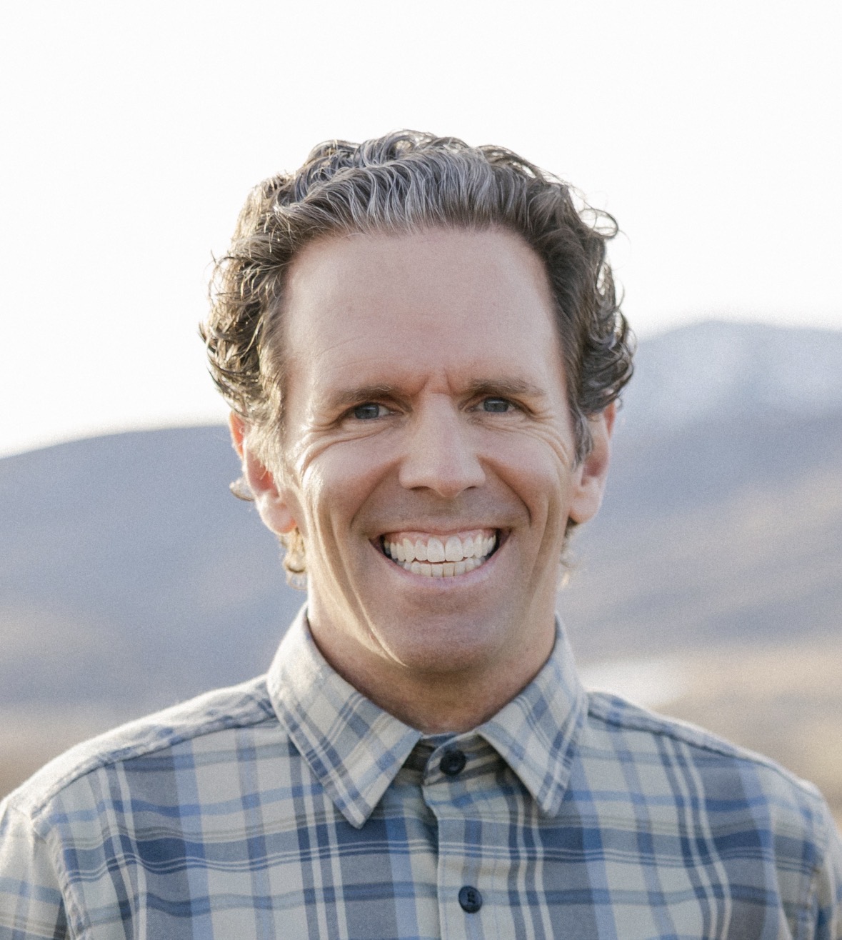 Smiling man with curly hair wearing a blue-and-gray plaid button-down shirt, standing outdoors with mountains softly blurred in the background.