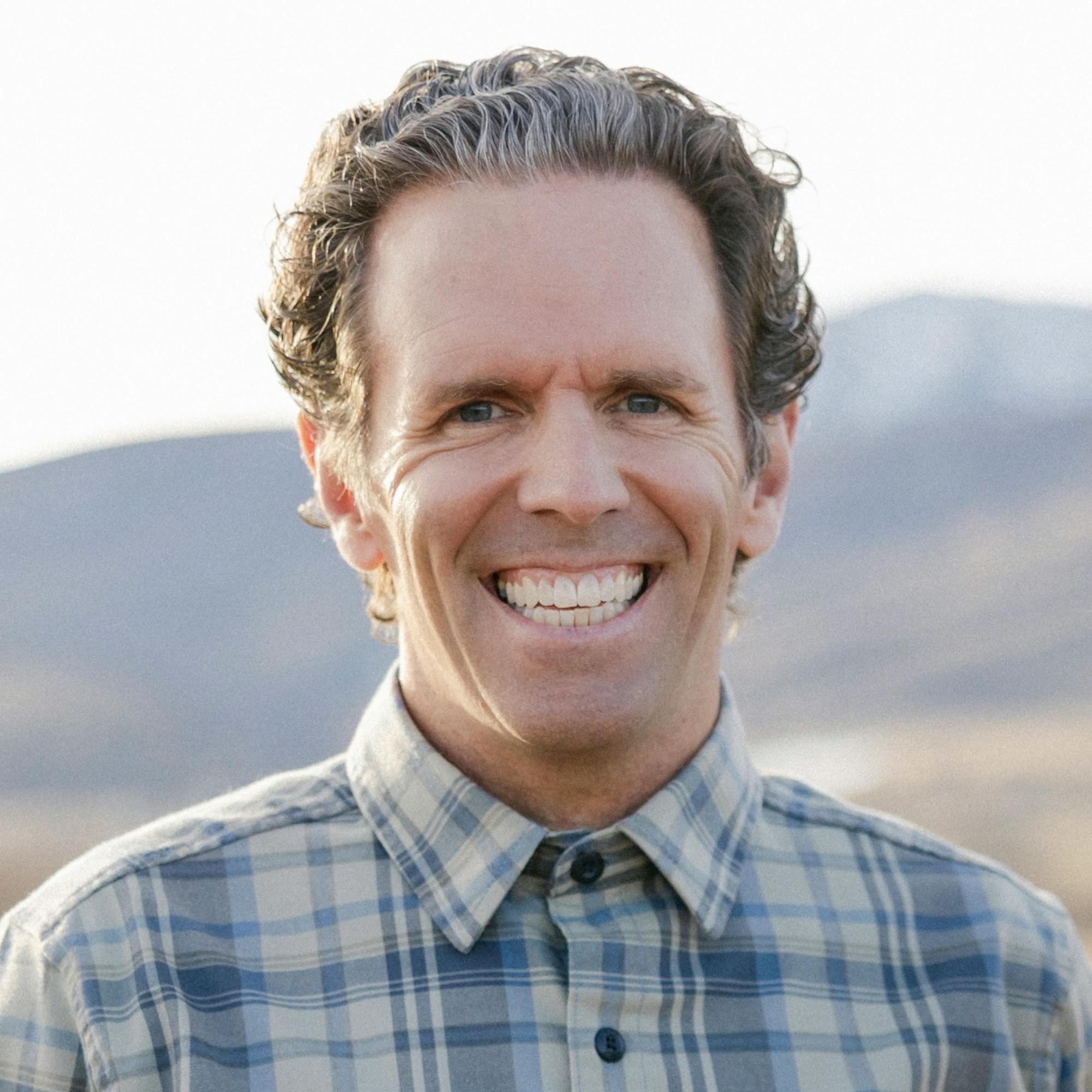 Smiling man with curly hair wearing a blue-and-gray plaid button-down shirt, standing outdoors with mountains softly blurred in the background.