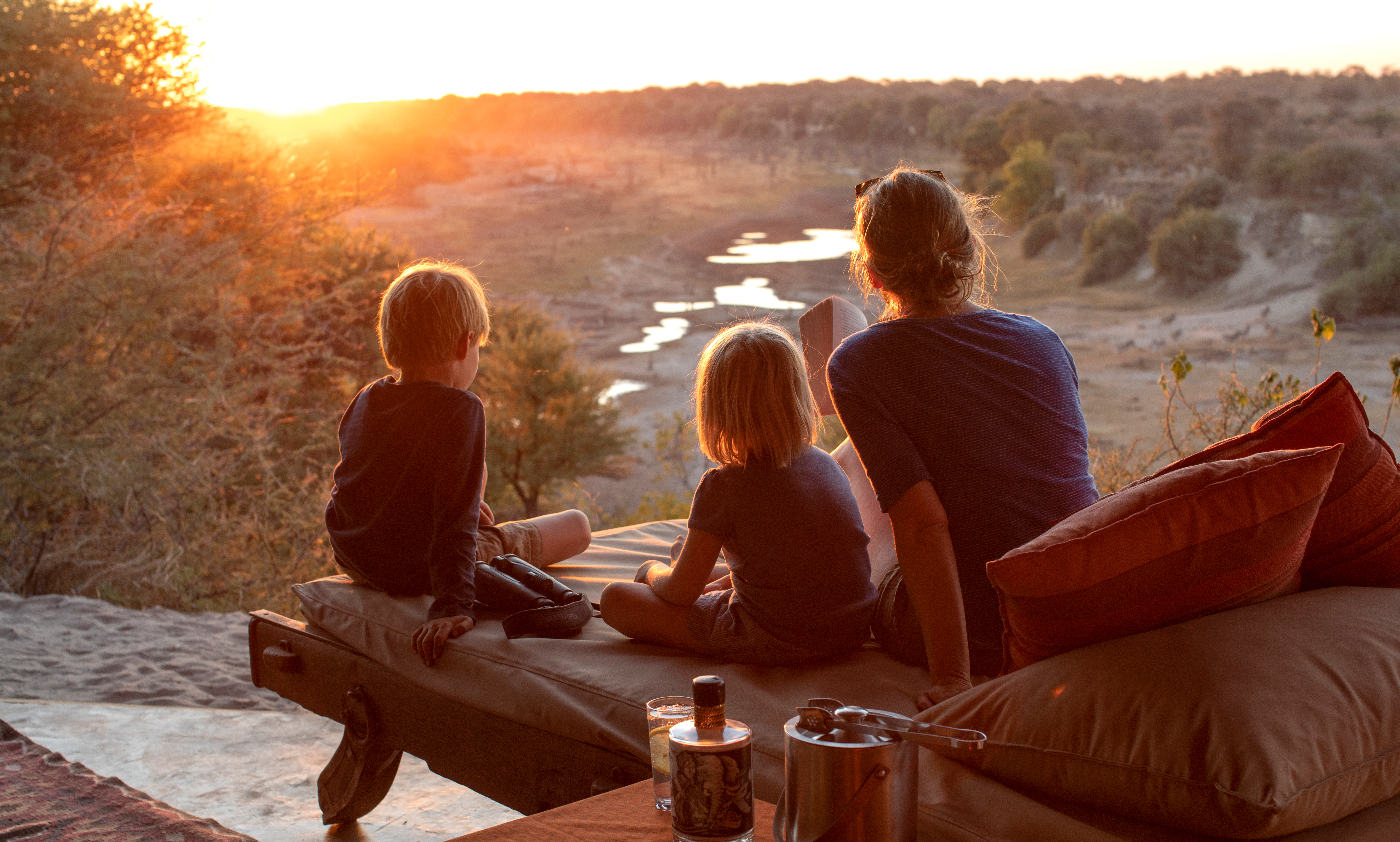 A family sits on a riverbank platform, watching the sun set over still water and distant trees as evening light fades.