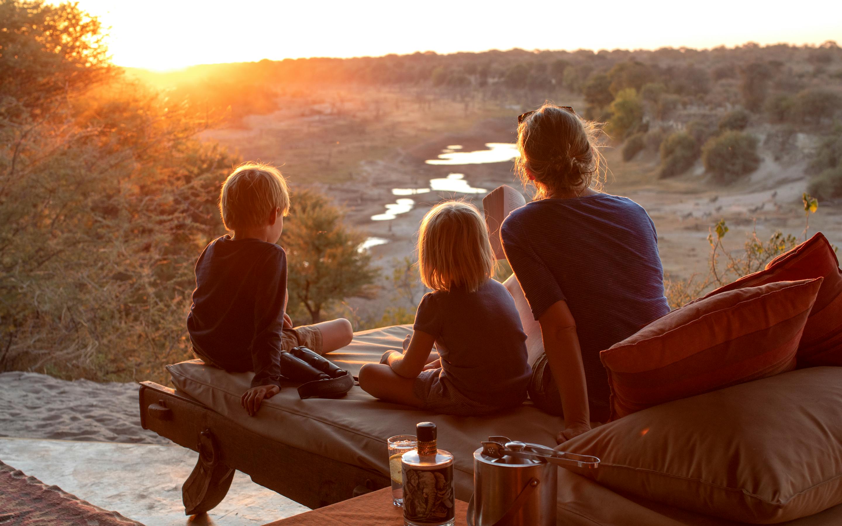 A family sits on a riverbank platform, watching the sun set over still water and distant trees as evening light fades.