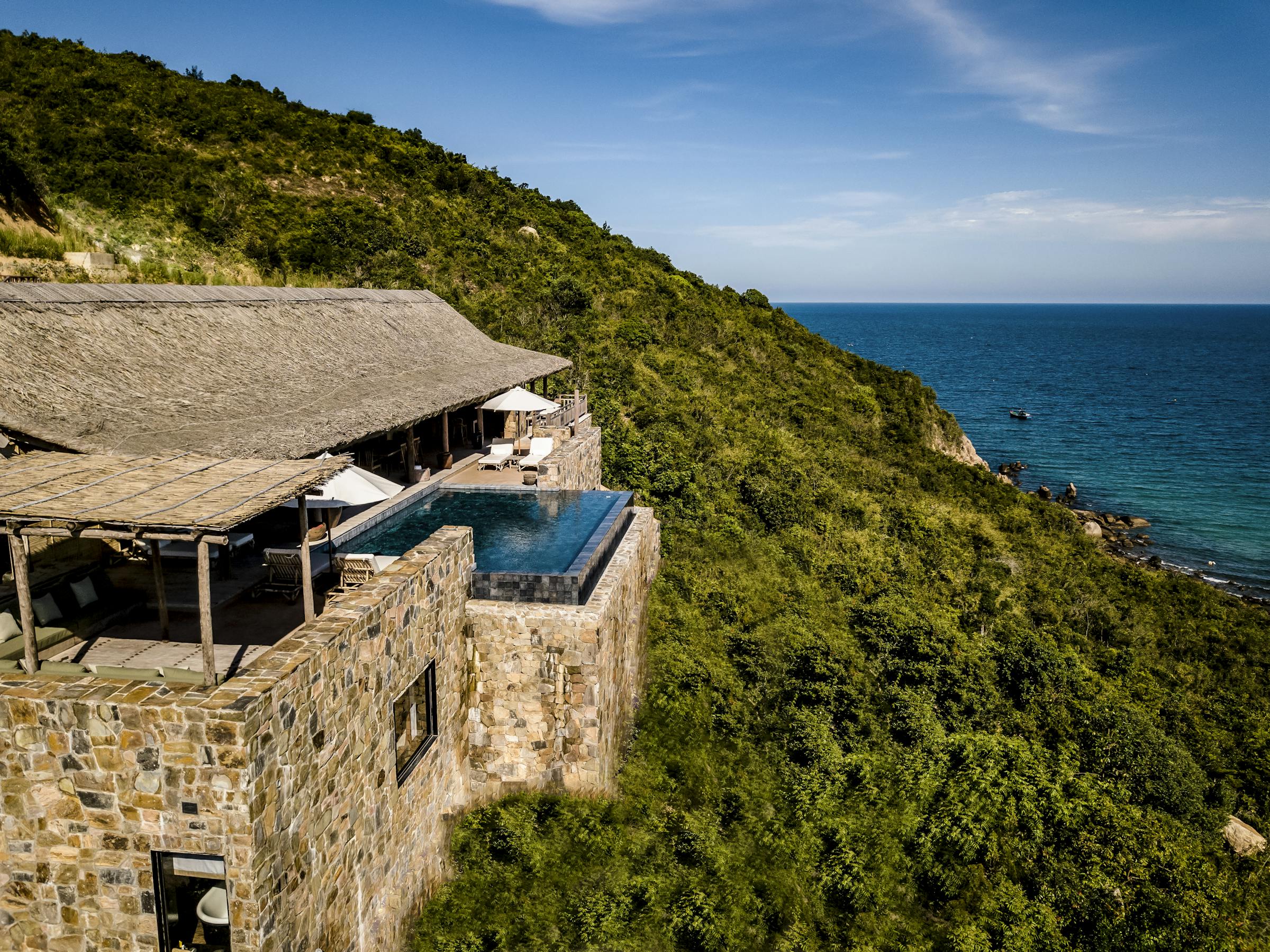 Infinity pool beside a cliffside villa overlooks the ocean, with lounge seating and a stone terrace under soft daylight.