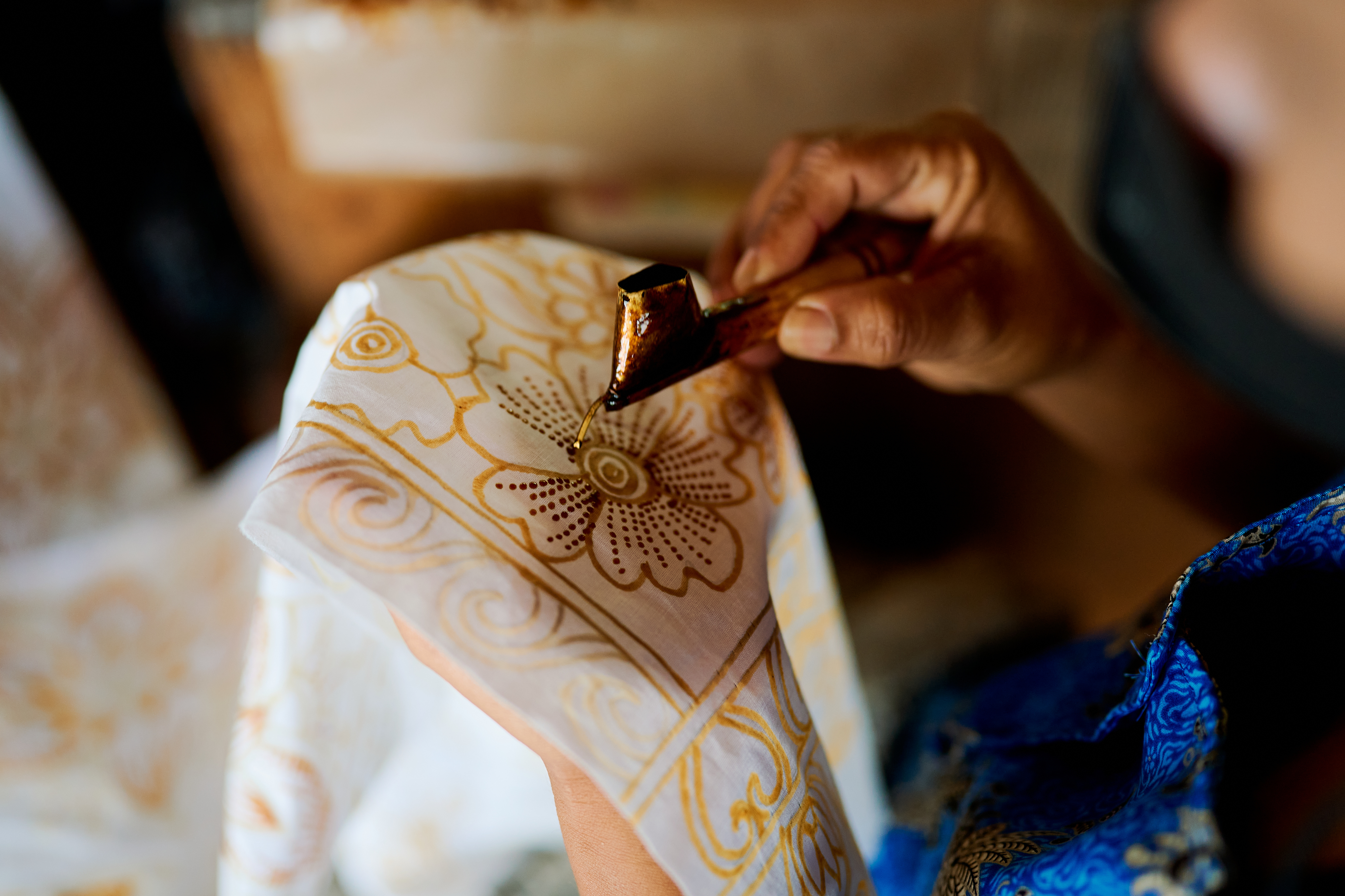 Hands draw a detailed batik pattern on fabric, with wax lines forming floral shapes on white cloth under soft daylight.