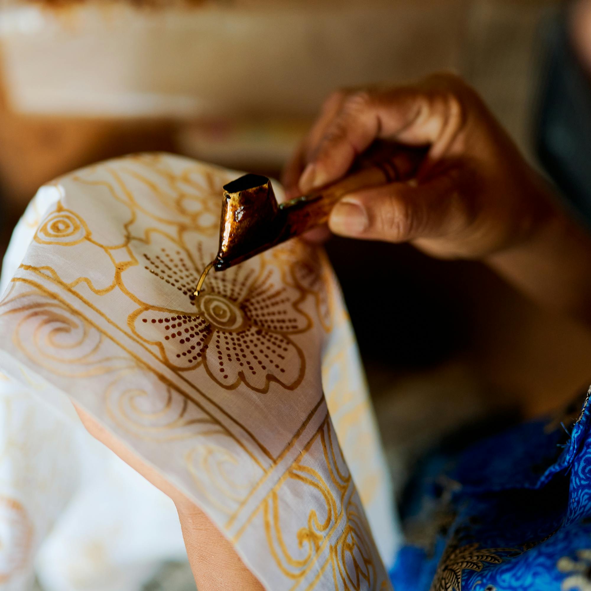 Hands draw a detailed batik pattern on fabric, with wax lines forming floral shapes on white cloth under soft daylight.