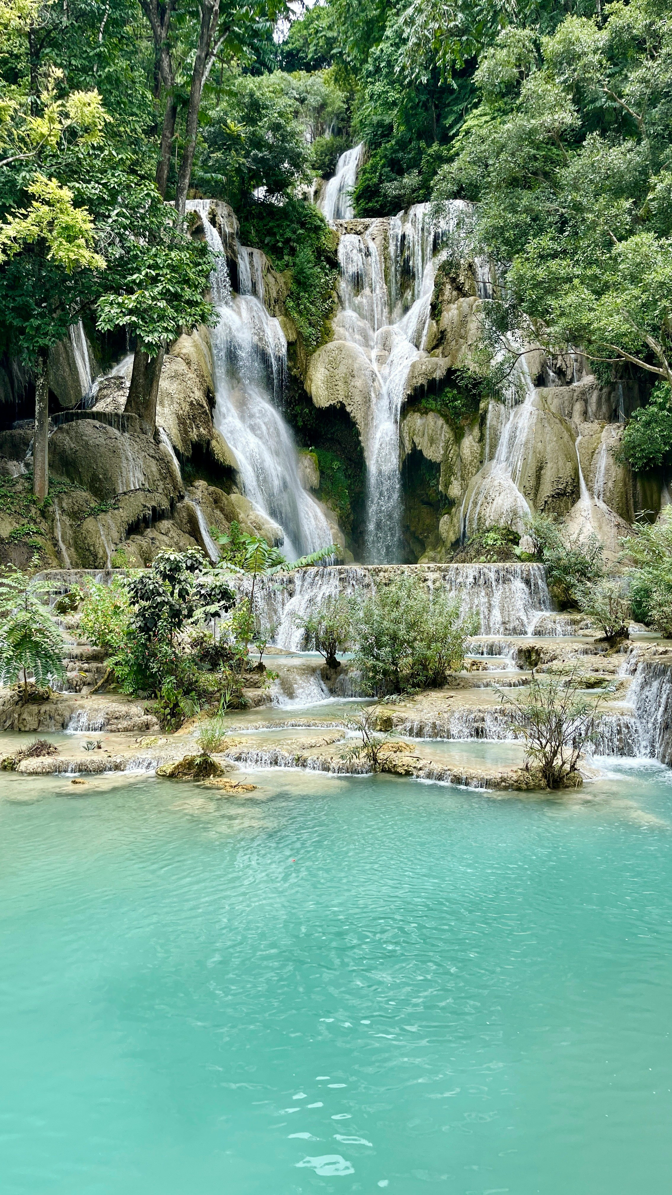 Waterfall cascades into turquoise pools, with mossy rocks and lush greenery surrounding the scene under soft daylight.