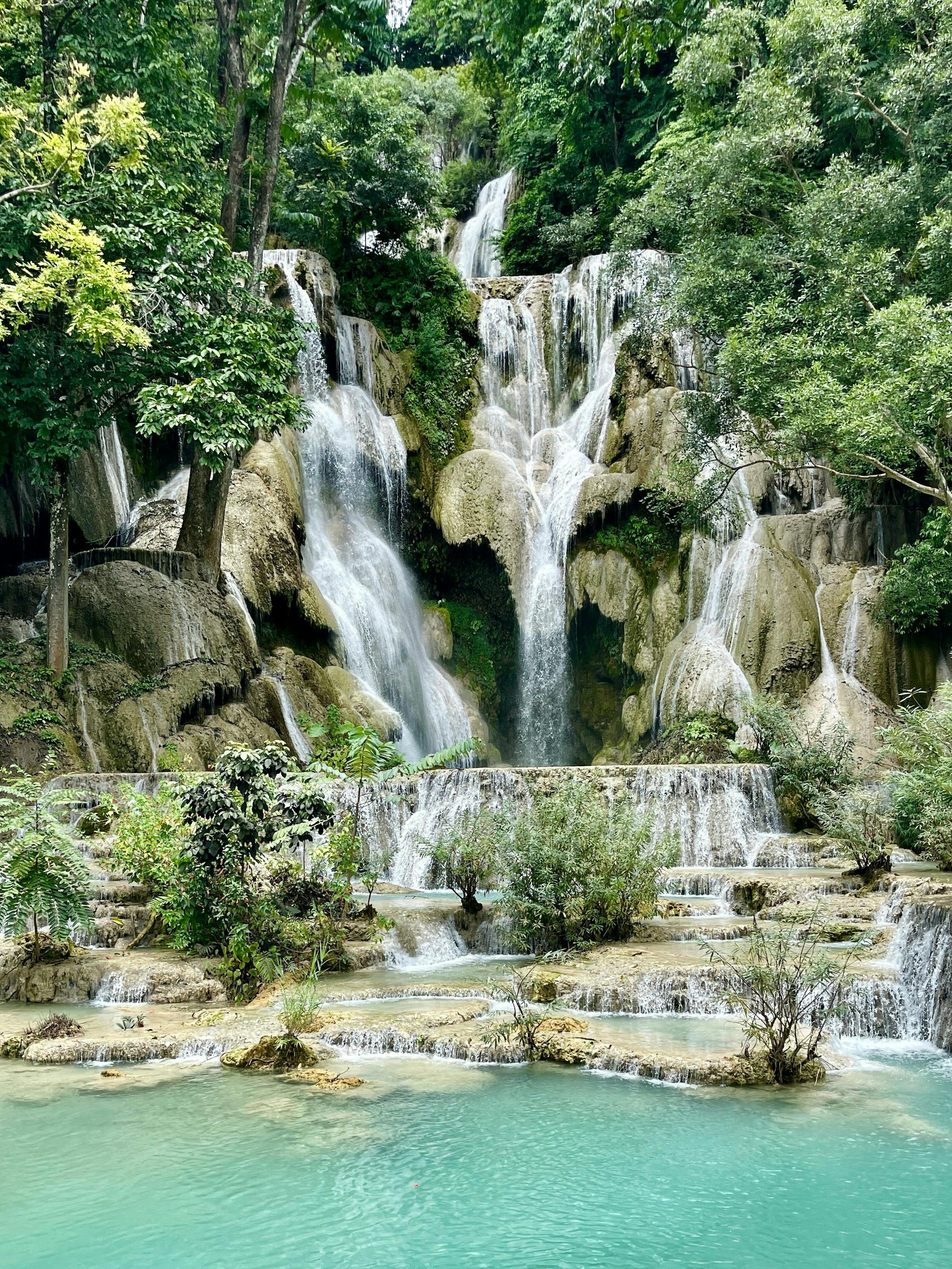 Waterfall cascades into turquoise pools, with mossy rocks and lush greenery surrounding the scene under soft daylight.