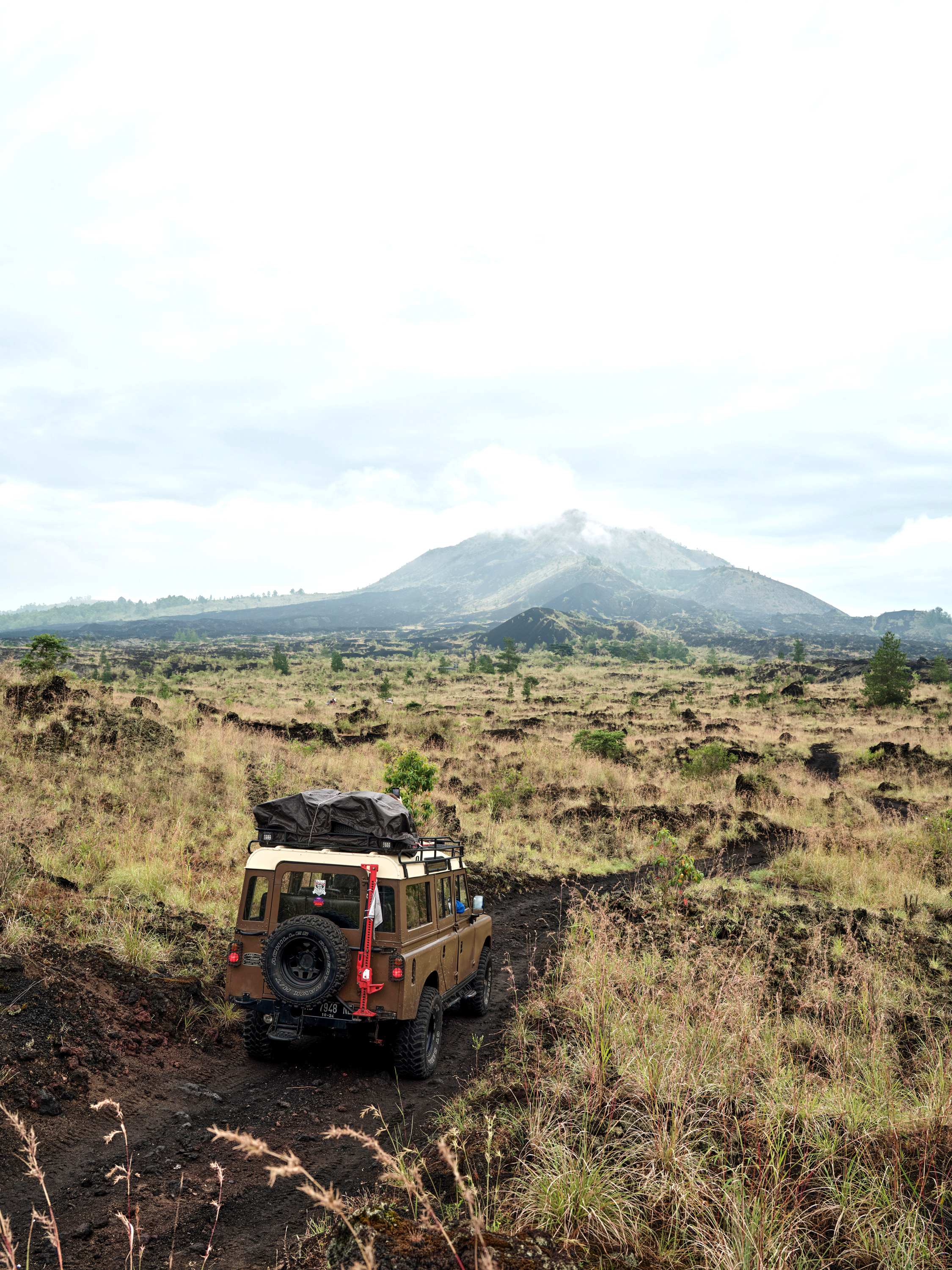 Red jeep drives along a rough track through open landscape, with mountains and a broad sky in the distance.