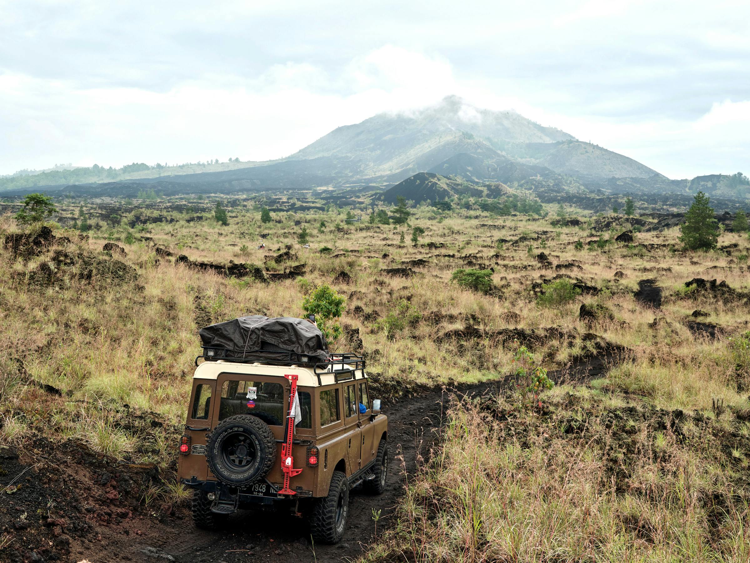 Red jeep drives along a rough track through open landscape, with mountains and a broad sky in the distance.