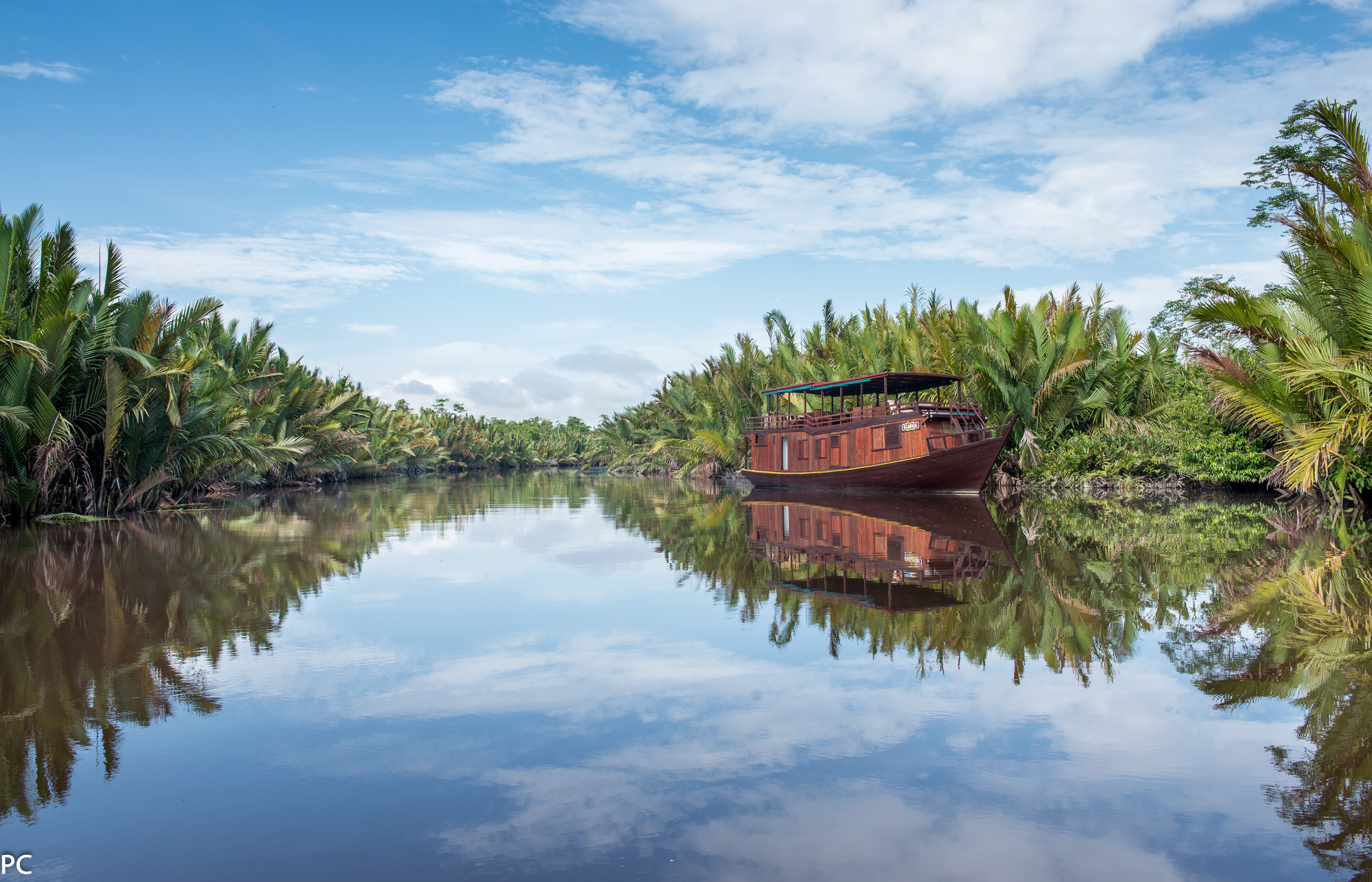 Wooden riverboat glides along a still river, with jungle trees reflected on the water in calm light under soft daylight.