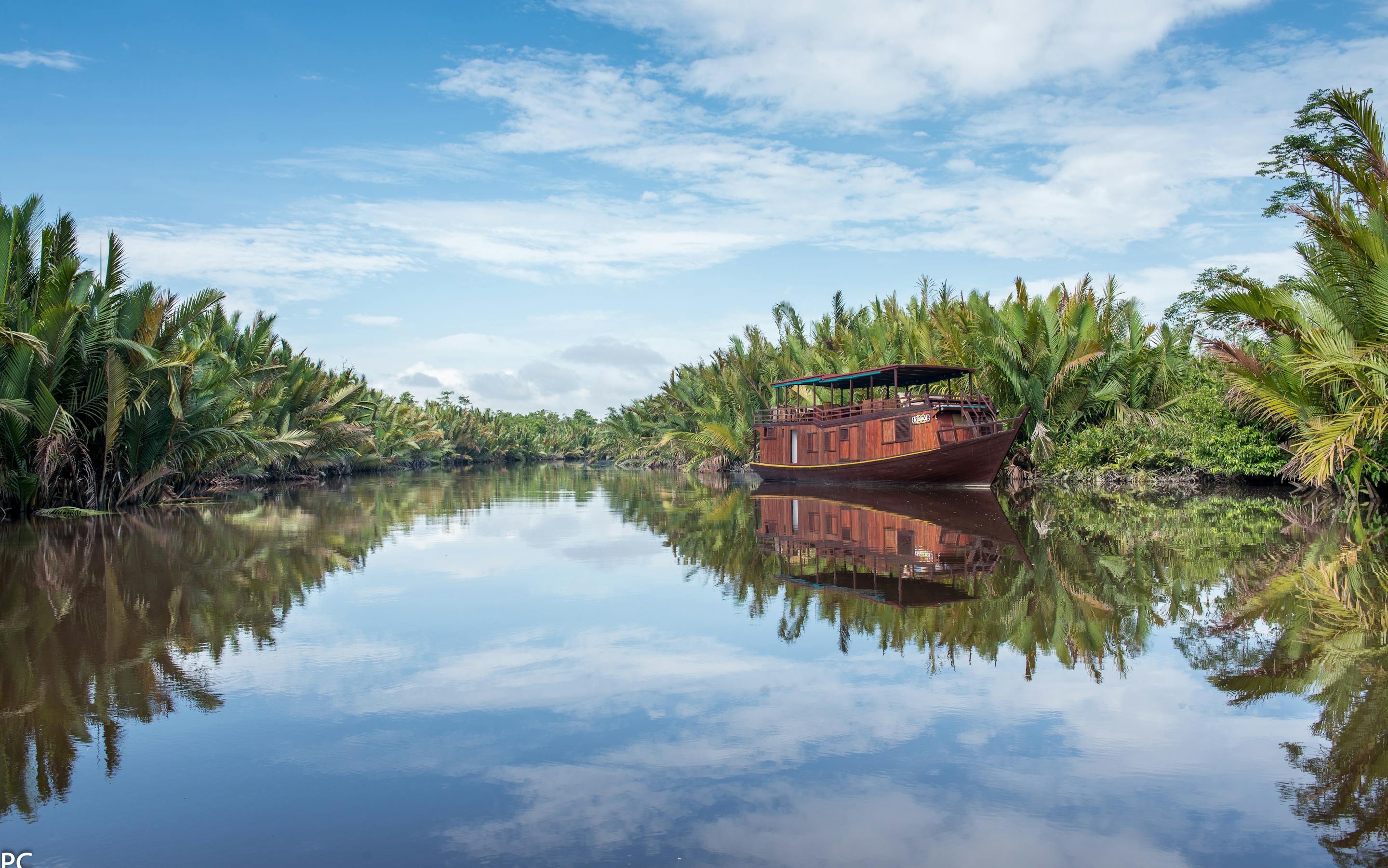 Wooden riverboat glides along a still river, with jungle trees reflected on the water in calm light under soft daylight.