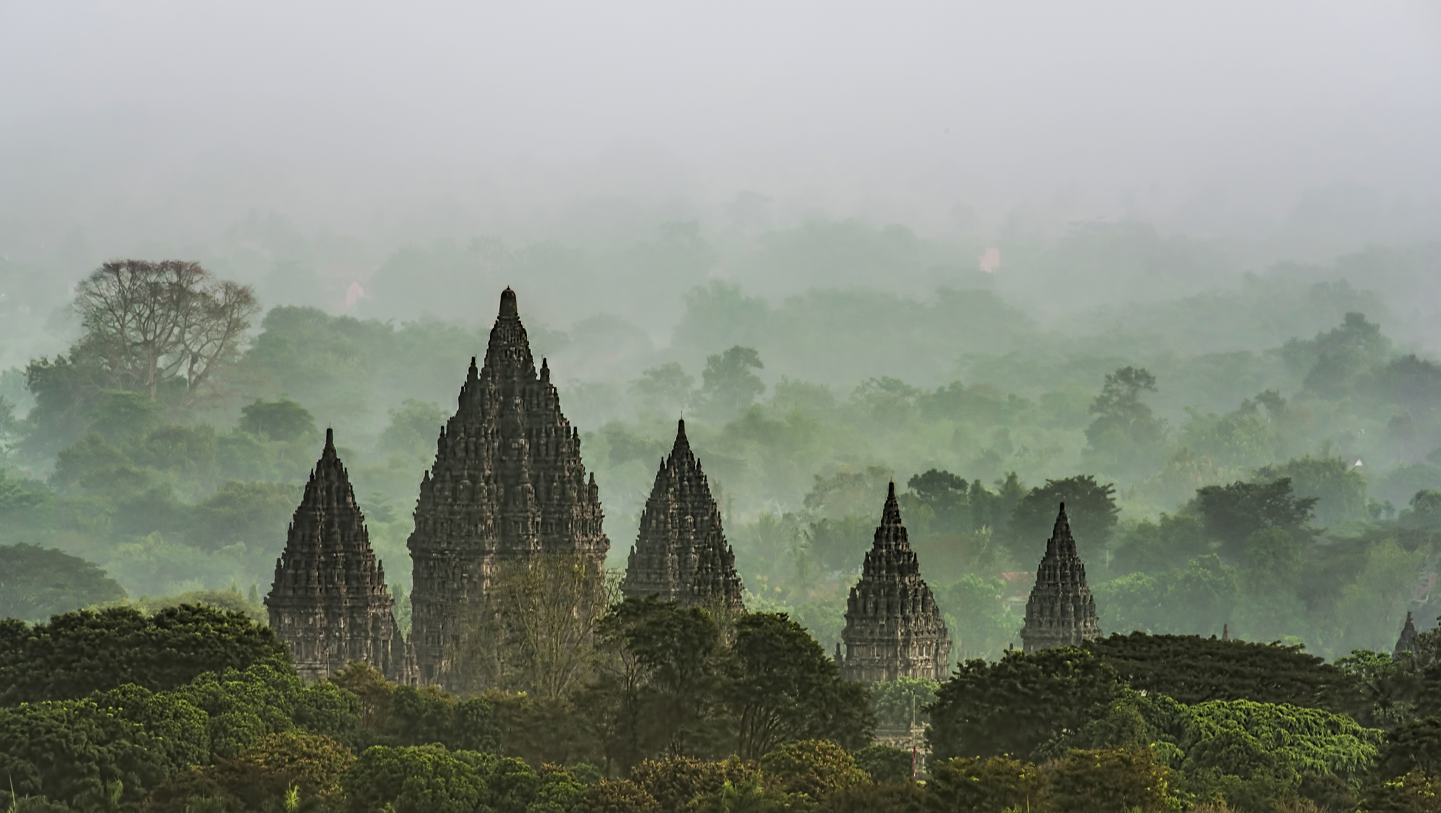 Ancient temple spires rise through morning mist, with soft light and layered silhouettes in the haze under soft daylight.