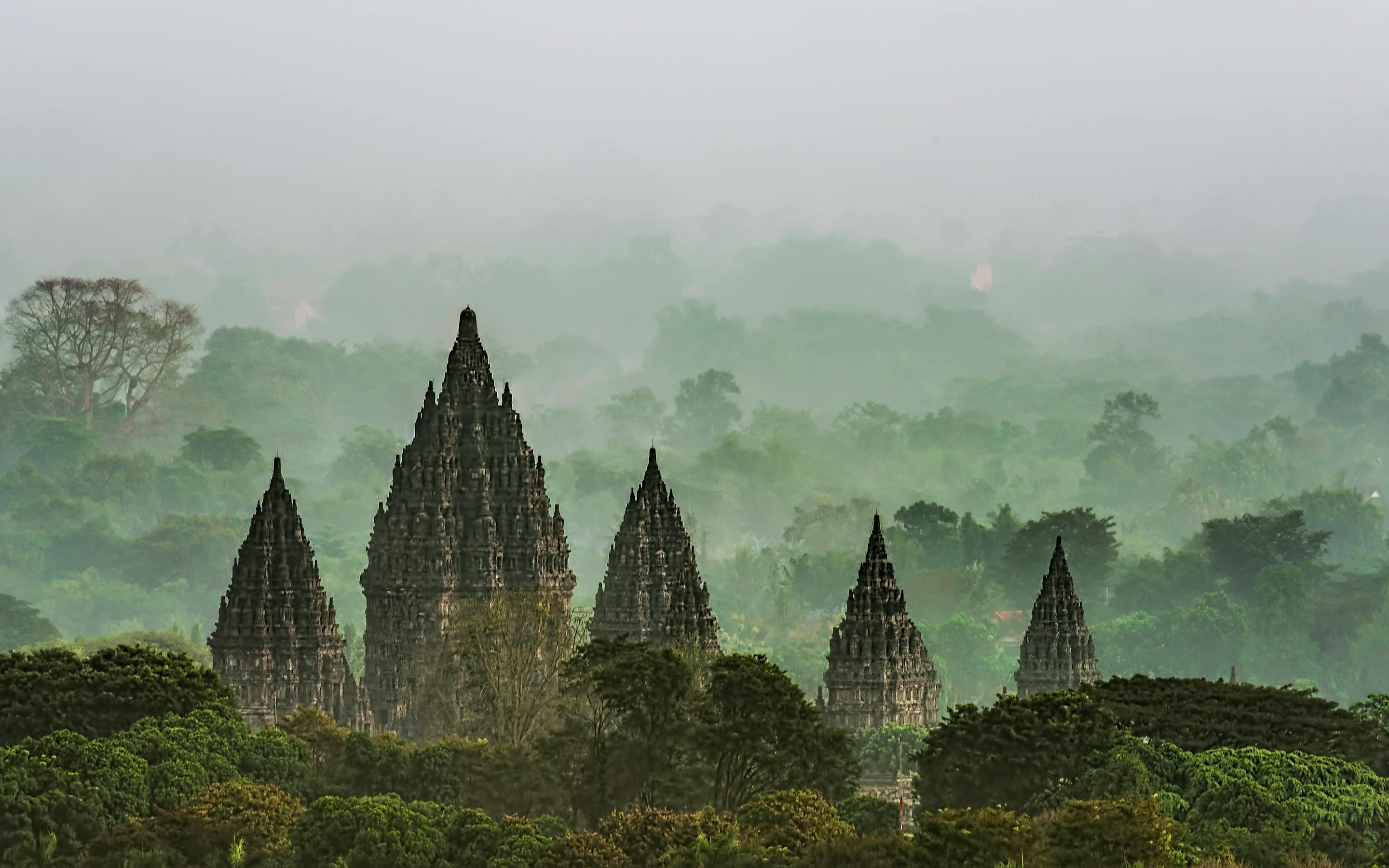 Ancient temple spires rise through morning mist, with soft light and layered silhouettes in the haze under soft daylight.