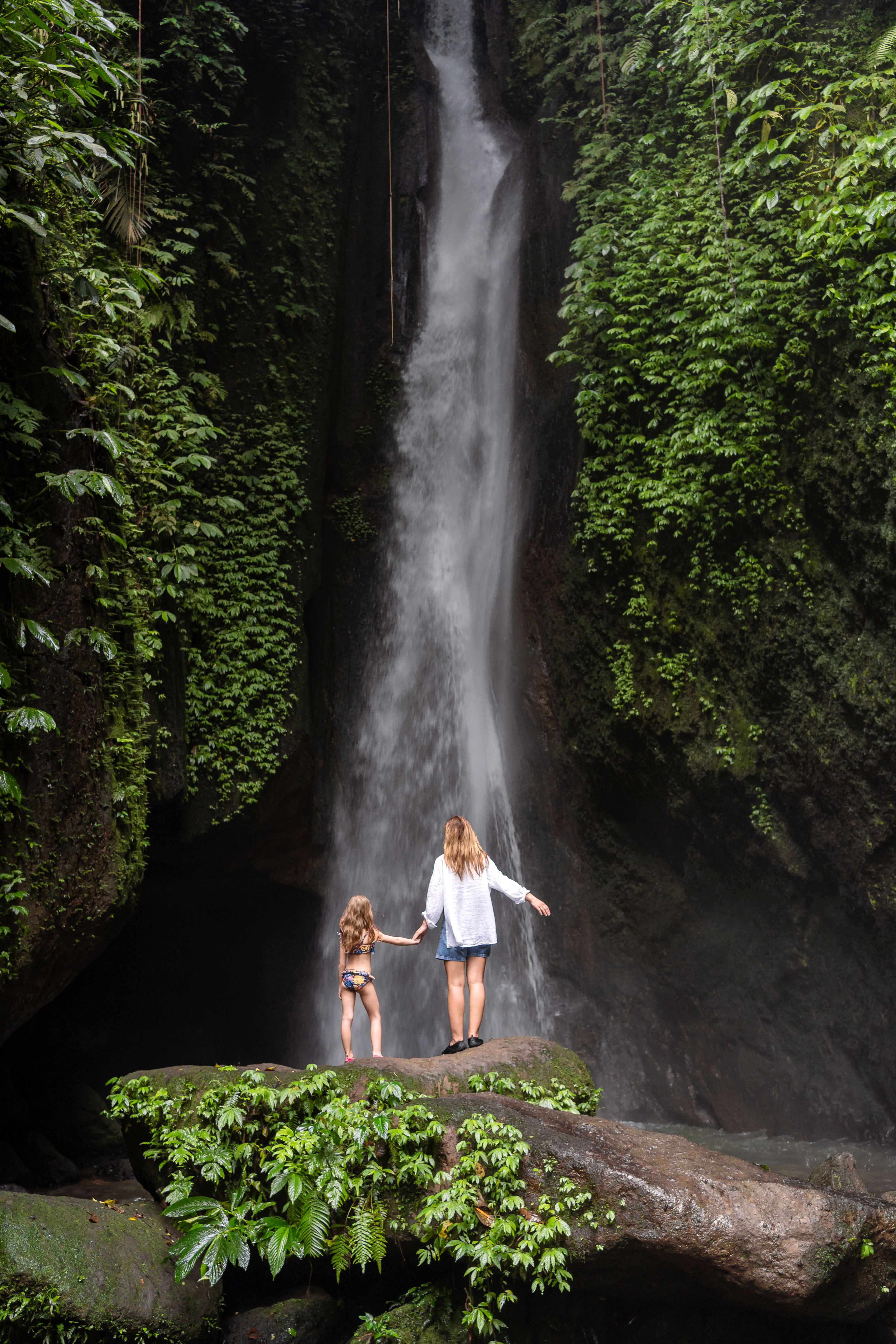 Mother and child stand beneath a tall waterfall in dense forest, with mist rising and ferns along wet rock walls.