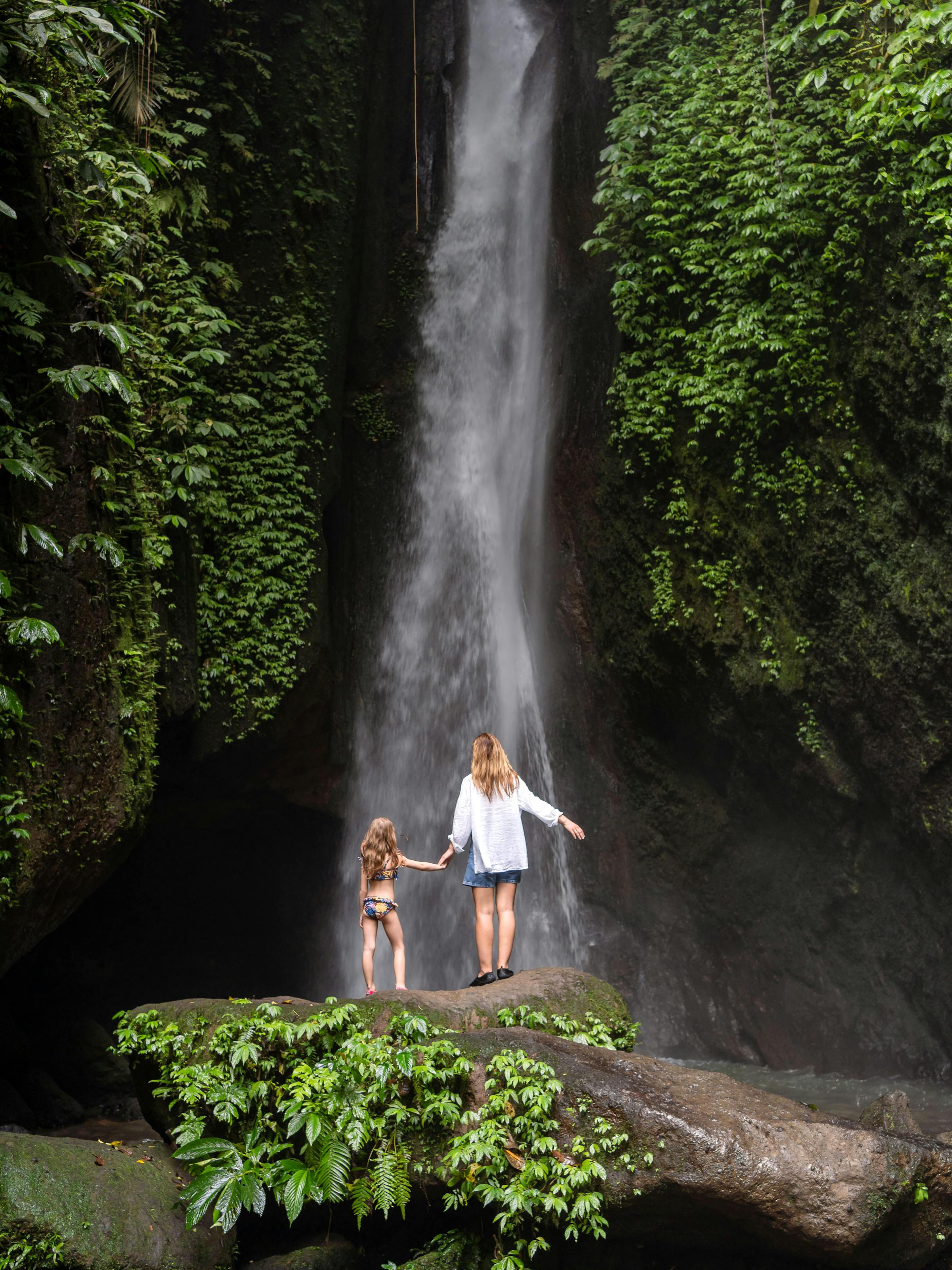 Mother and child stand beneath a tall waterfall in dense forest, with mist rising and ferns along wet rock walls.