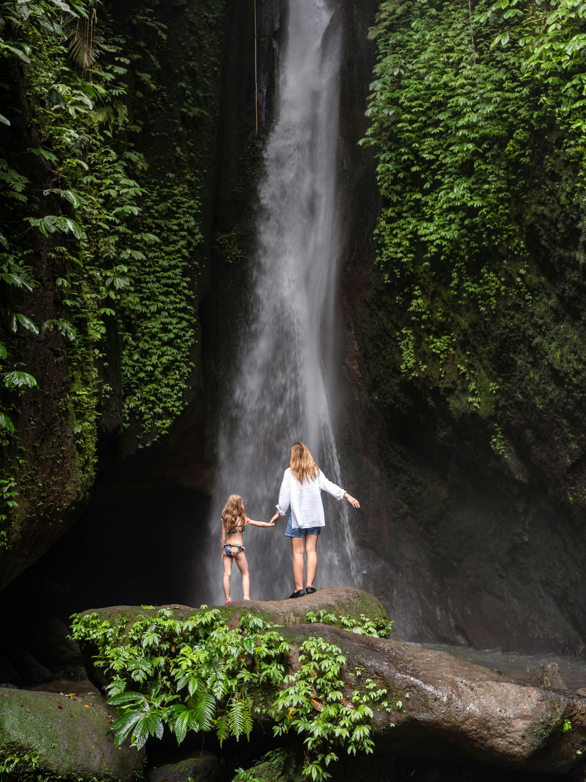 Mother and child stand beneath a tall waterfall in dense forest, with mist rising and ferns along wet rock walls.