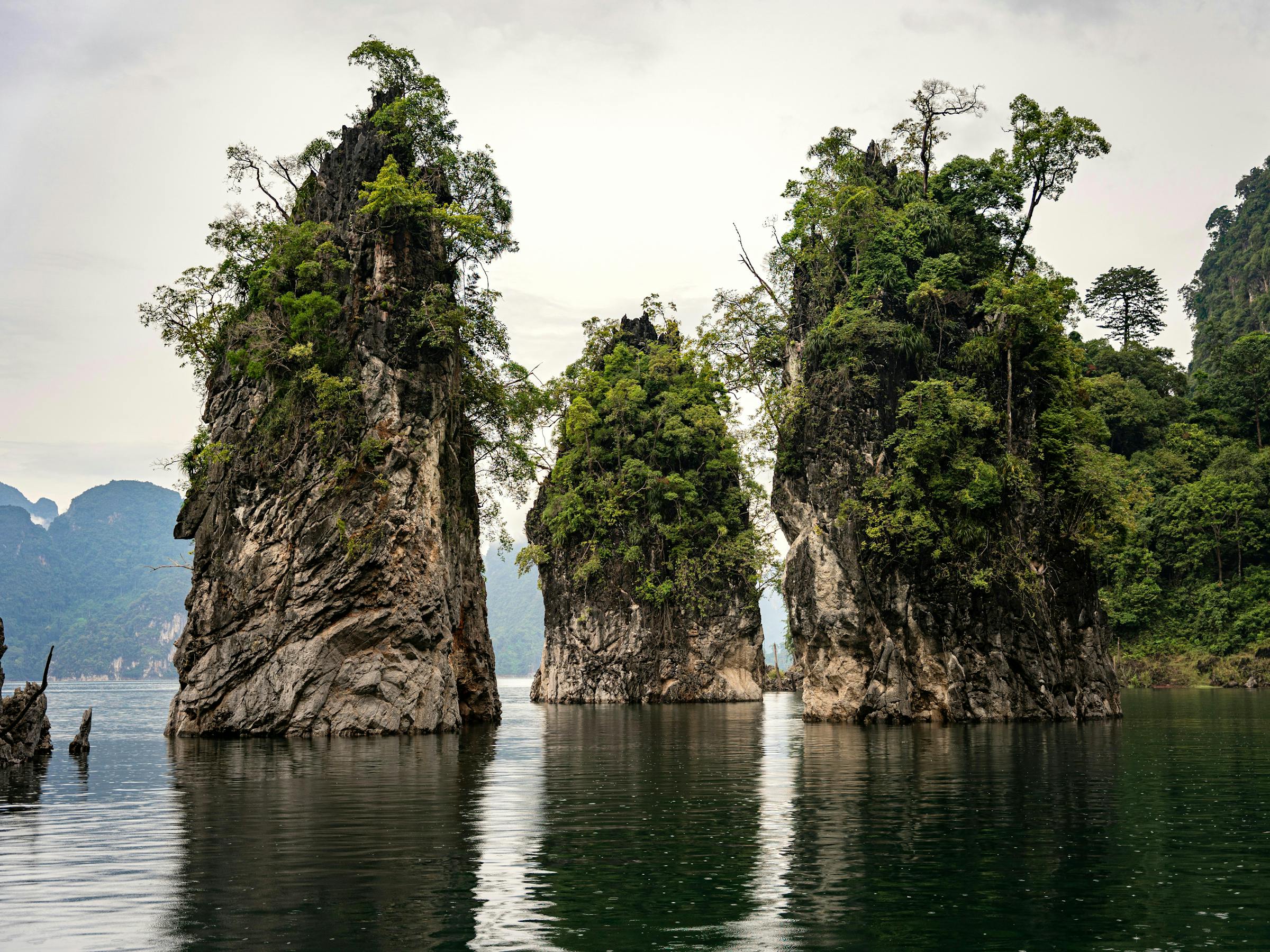 Tall limestone pillars rise from a calm lake, with green reflections and a gray sky above still water under soft daylight.