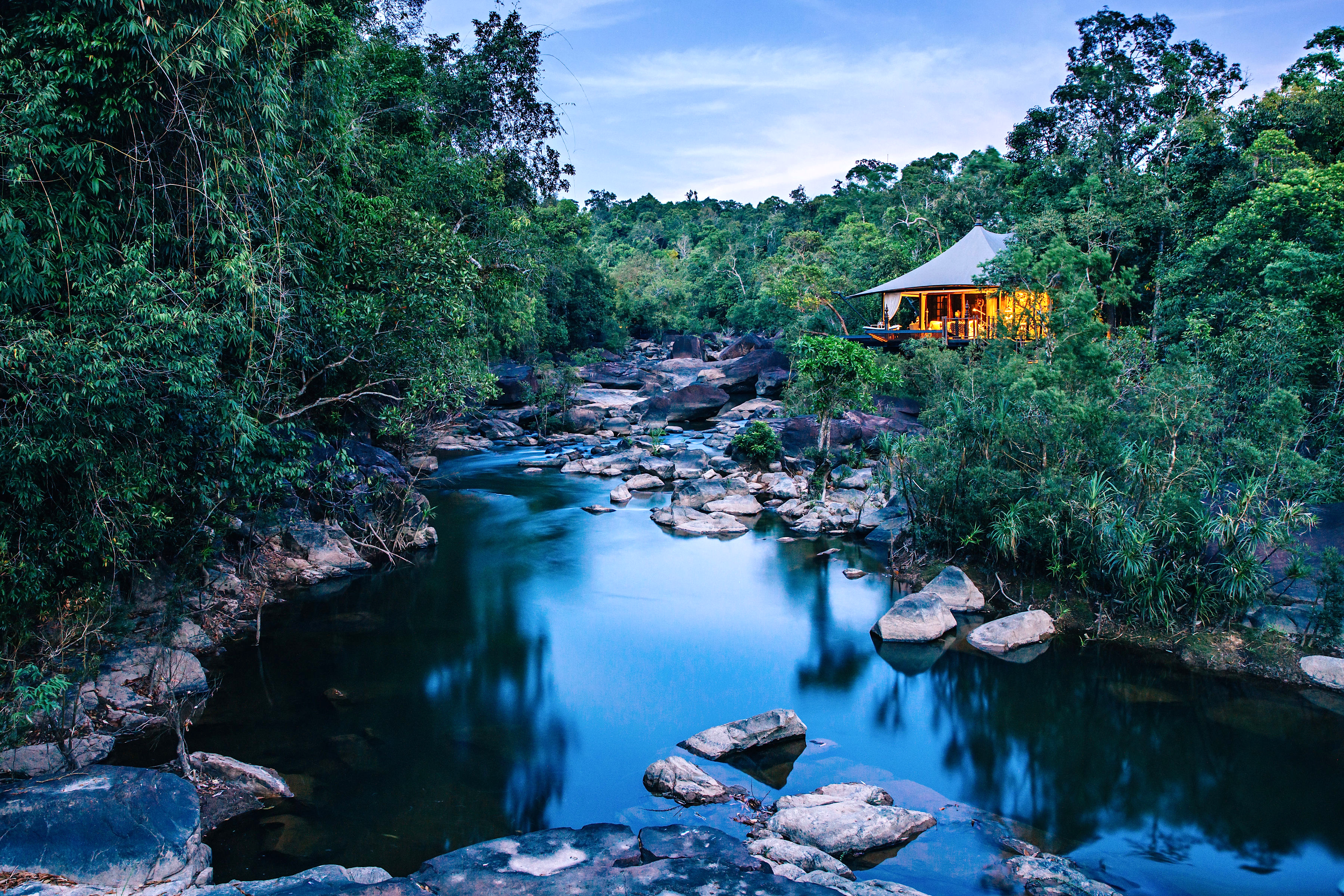 Jungle lodge tents sit beside a river pool, with smooth boulders and dense green foliage around the water.