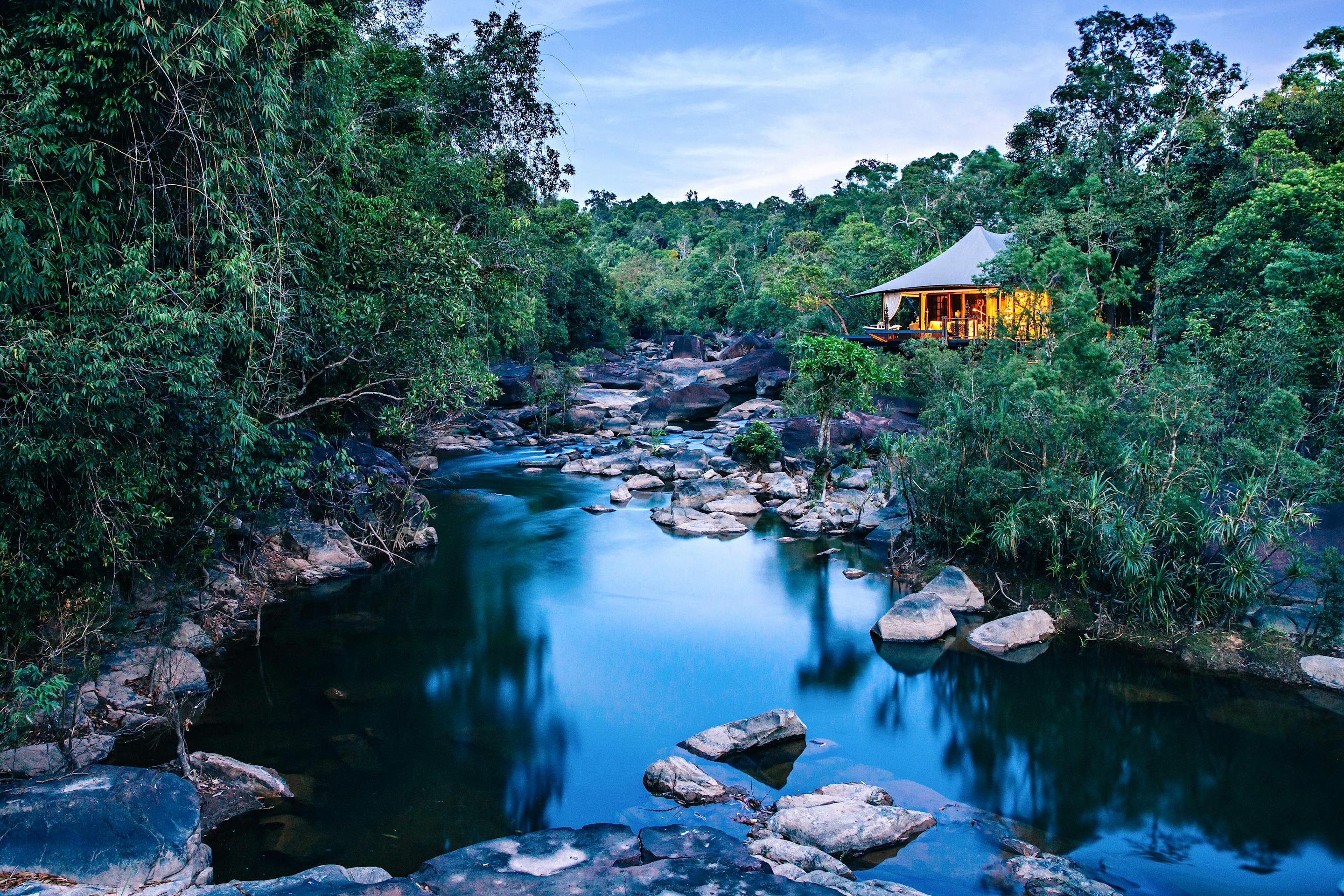 Jungle lodge tents sit beside a river pool, with smooth boulders and dense green foliage around the water.