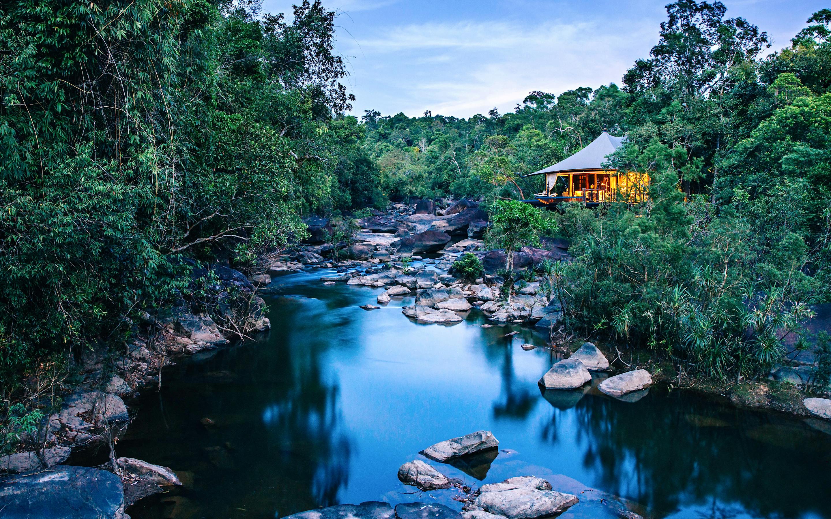Jungle lodge tents sit beside a river pool, with smooth boulders and dense green foliage around the water.