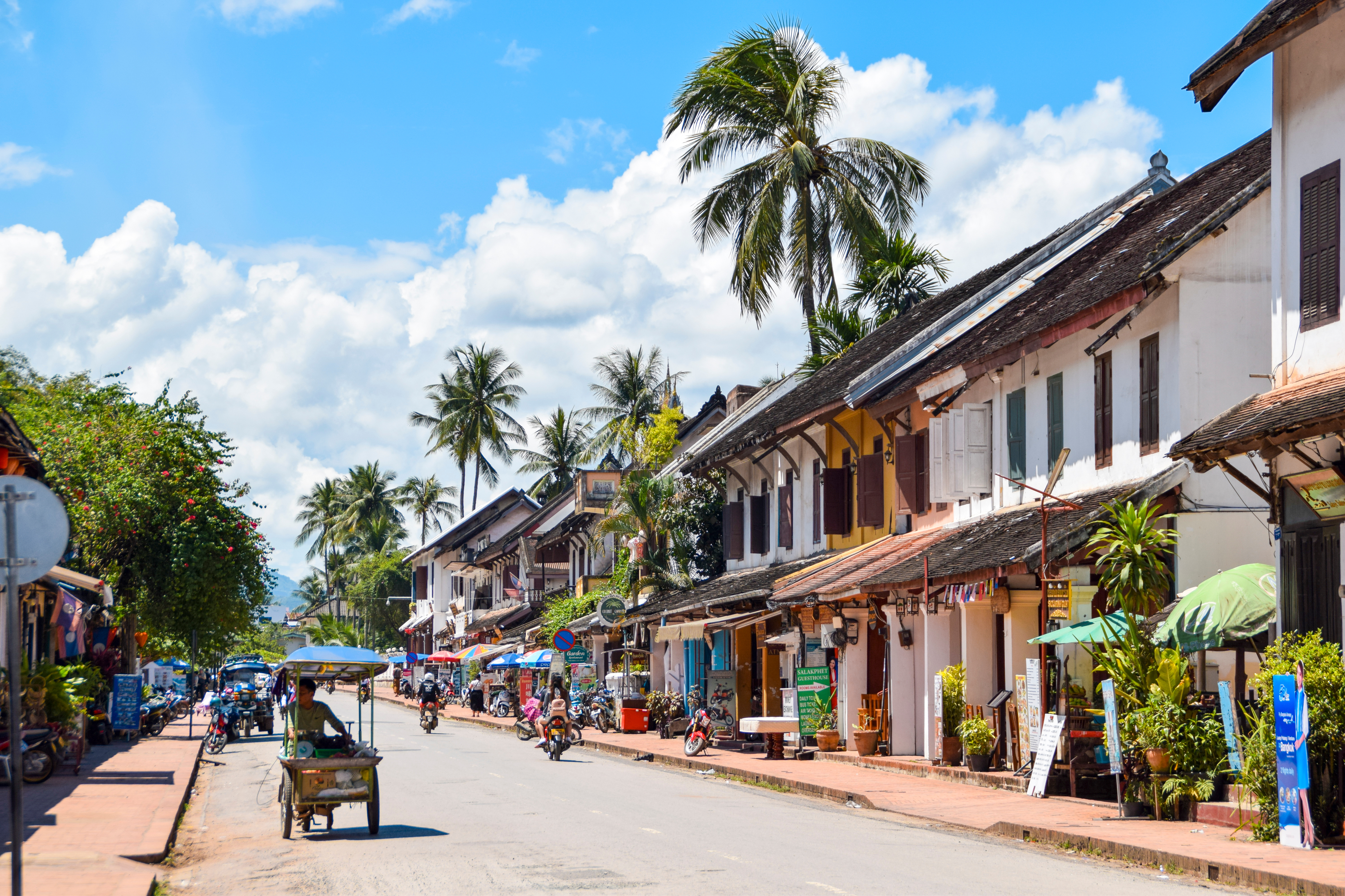 Sunlit street lined with low buildings and palm trees, with scooters and pedestrians in the distance under soft daylight.